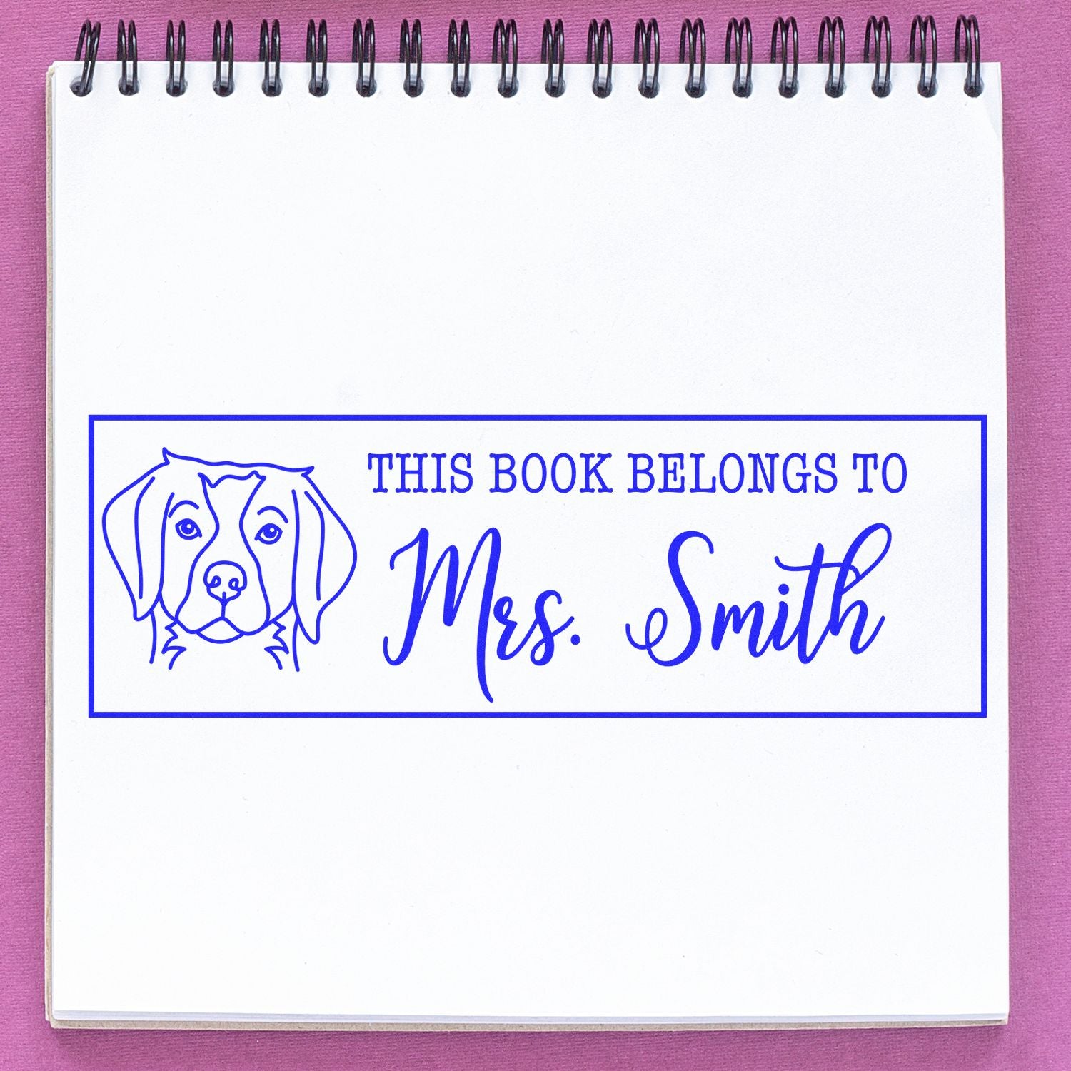 Blue custom book stamp with dog outline and This Book Belongs to Mrs. Smith on white notebook, pink background.