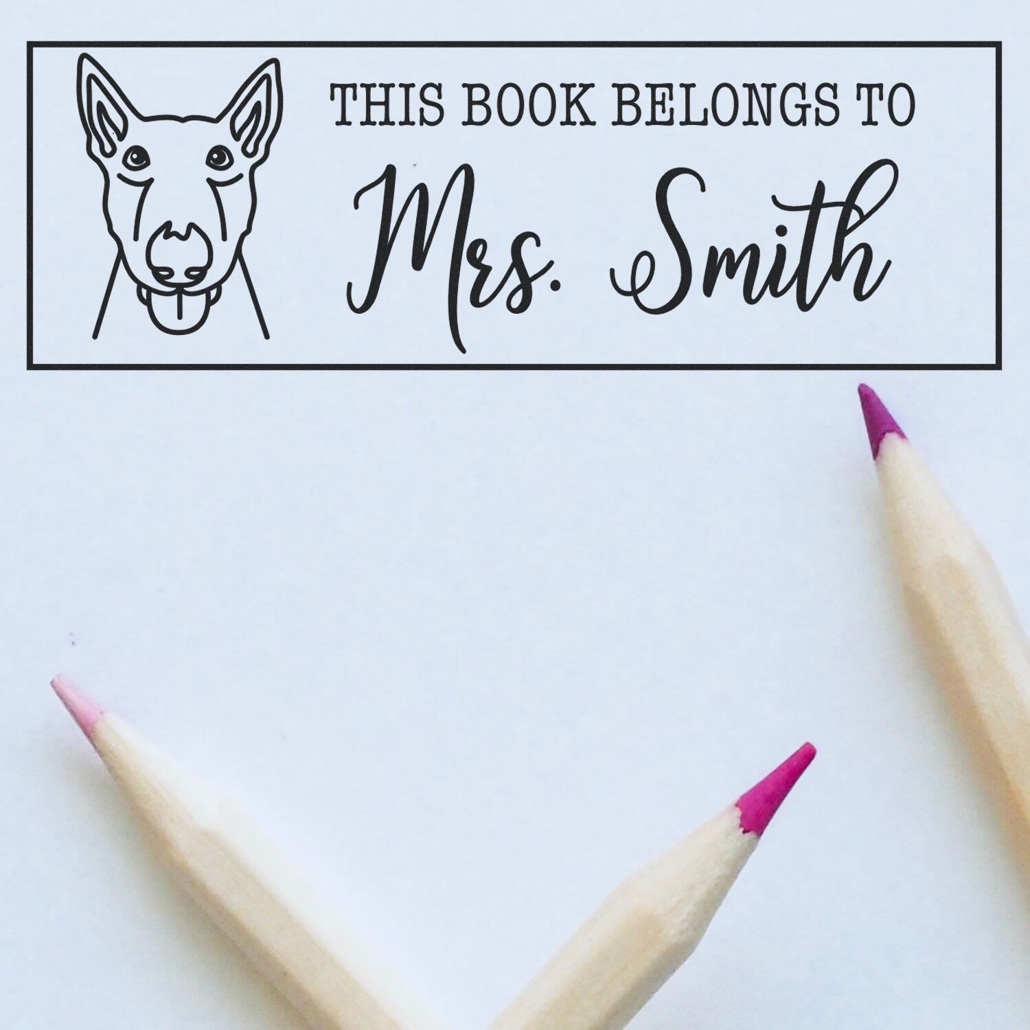 Custom black stamp and seal with dog outline, This Book Belongs to Mrs. Smith, on white paper with pink-tipped pencils.