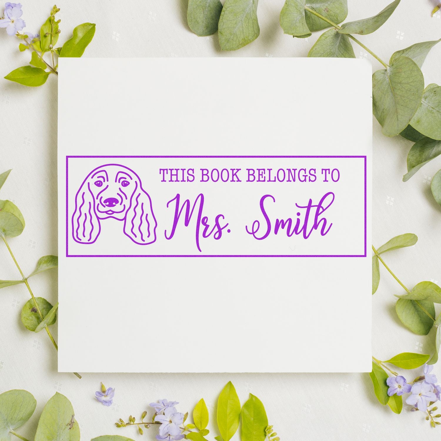 Purple custom book stamp and seal with dog outline, reading This Book Belongs To Mrs. Smith, on a white card with green leaves.