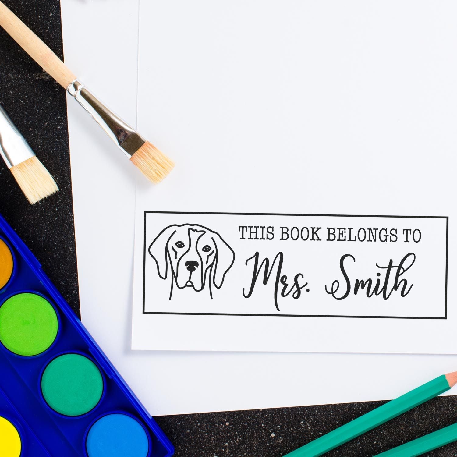 Custom black book stamp and seal for Mrs. Smith with dog illustration on white paper, beside paintbrushes and watercolor paints.