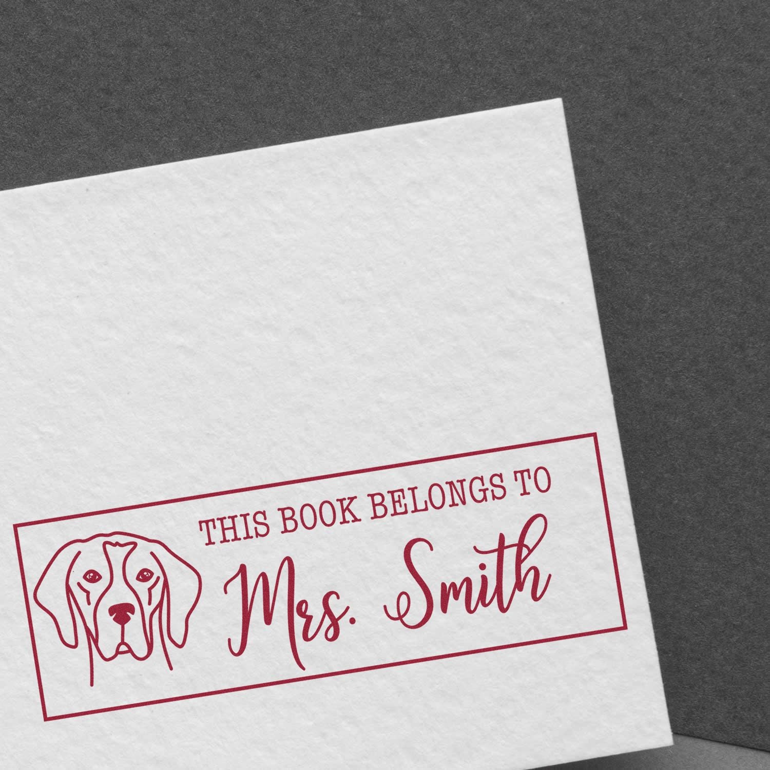 Red custom book stamp seal with dog outline and This book belongs to Mrs. Smith on white paper over dark gray background.