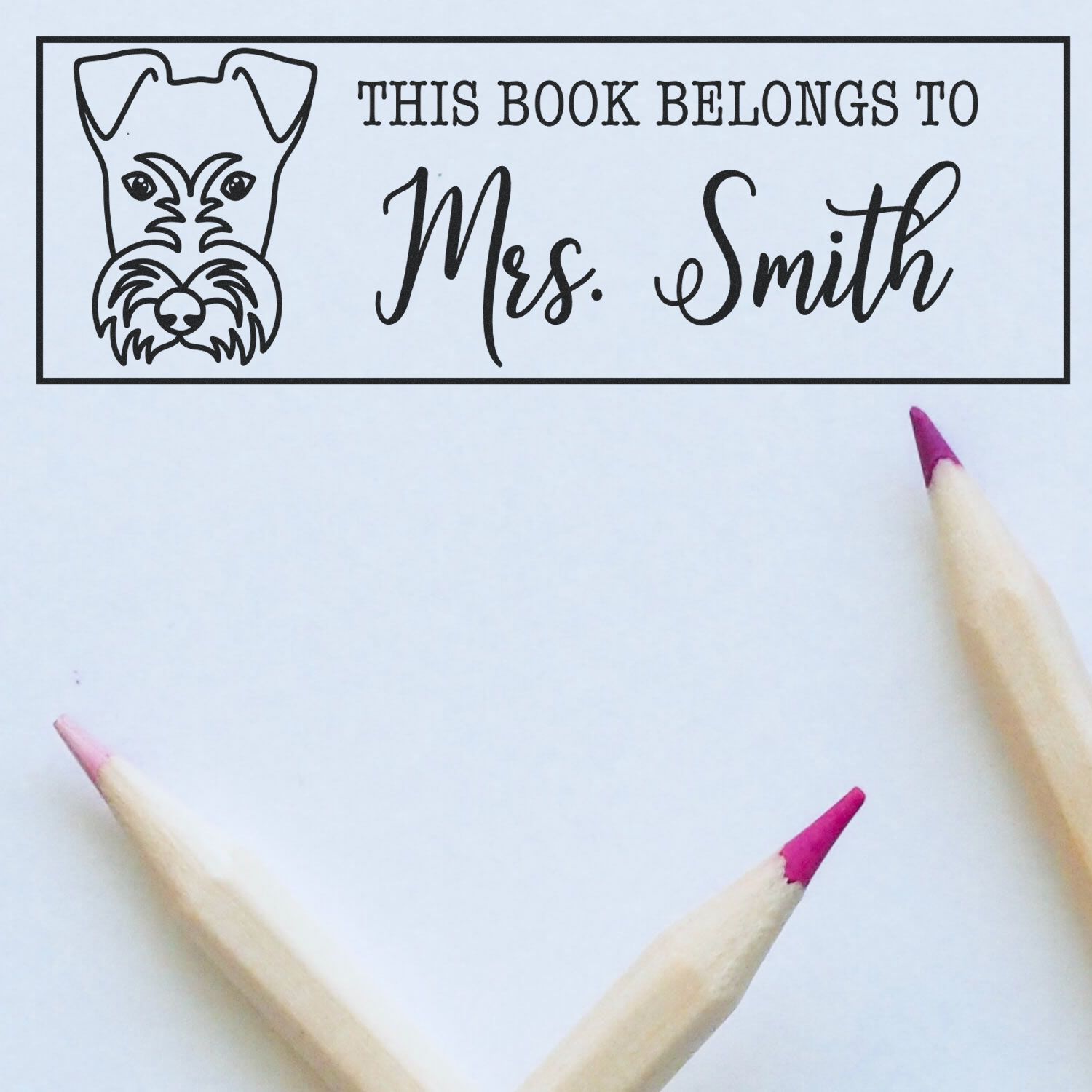 Black bookplate stamp with dog illustration and This Book Belongs to Mrs. Smith on white paper, shown with pink pencils.