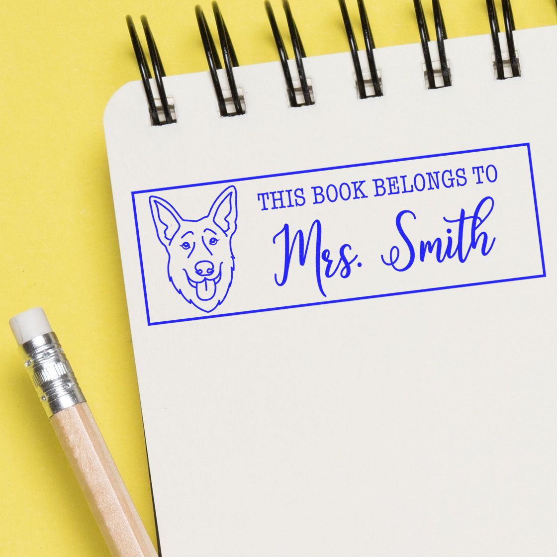 Blue custom book stamp with dog seal reading This Book Belongs to Mrs. Smith on white notebook, yellow background.