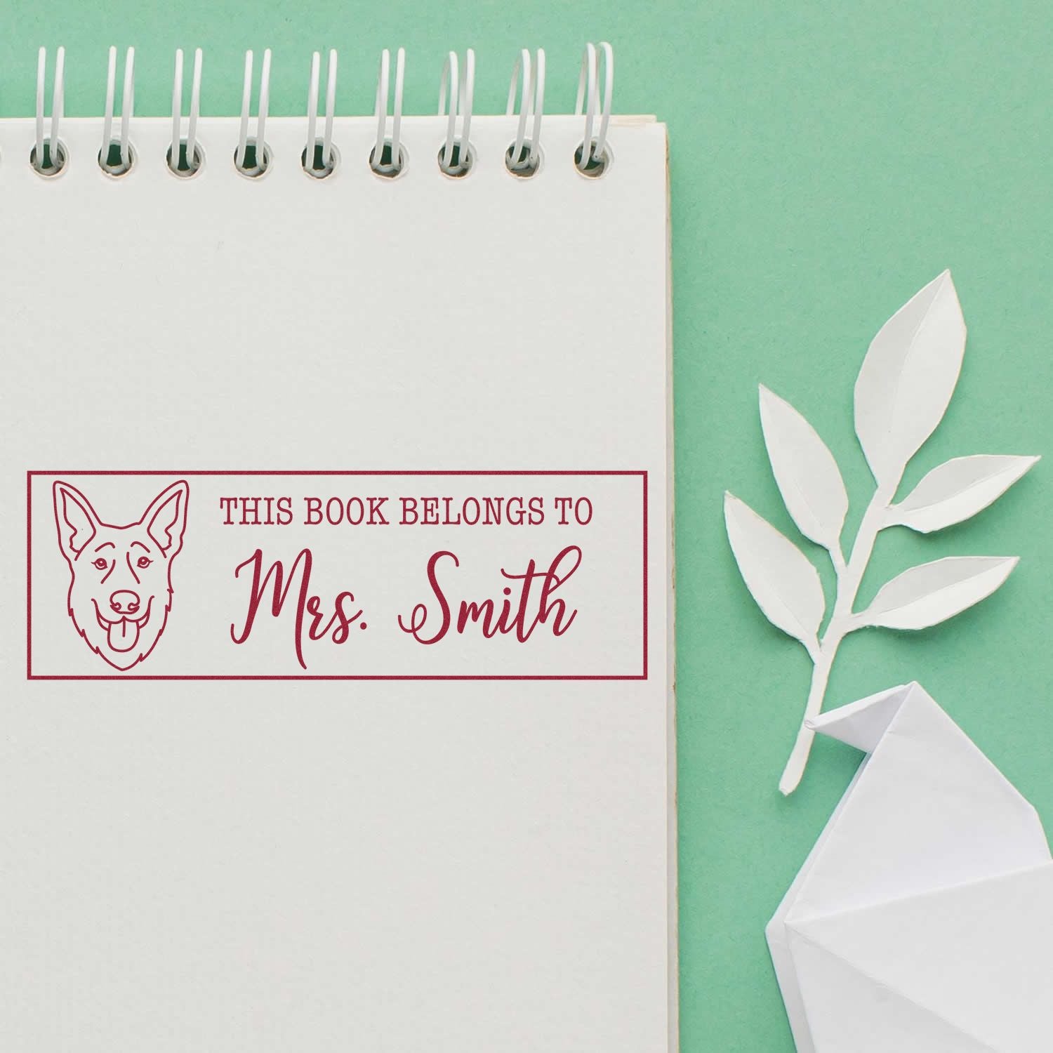 Custom red book stamp with dog icon and Mrs. Smith on white notebook, beside white paper leaf and envelope on mint green background.