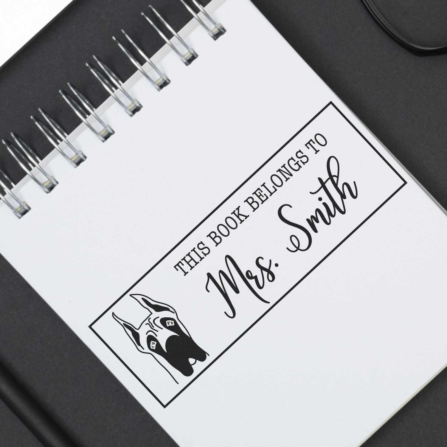 Black-and-white custom stamp and seal on a spiral notebook, reading This Book Belongs to Mrs. Smith with a dog illustration.