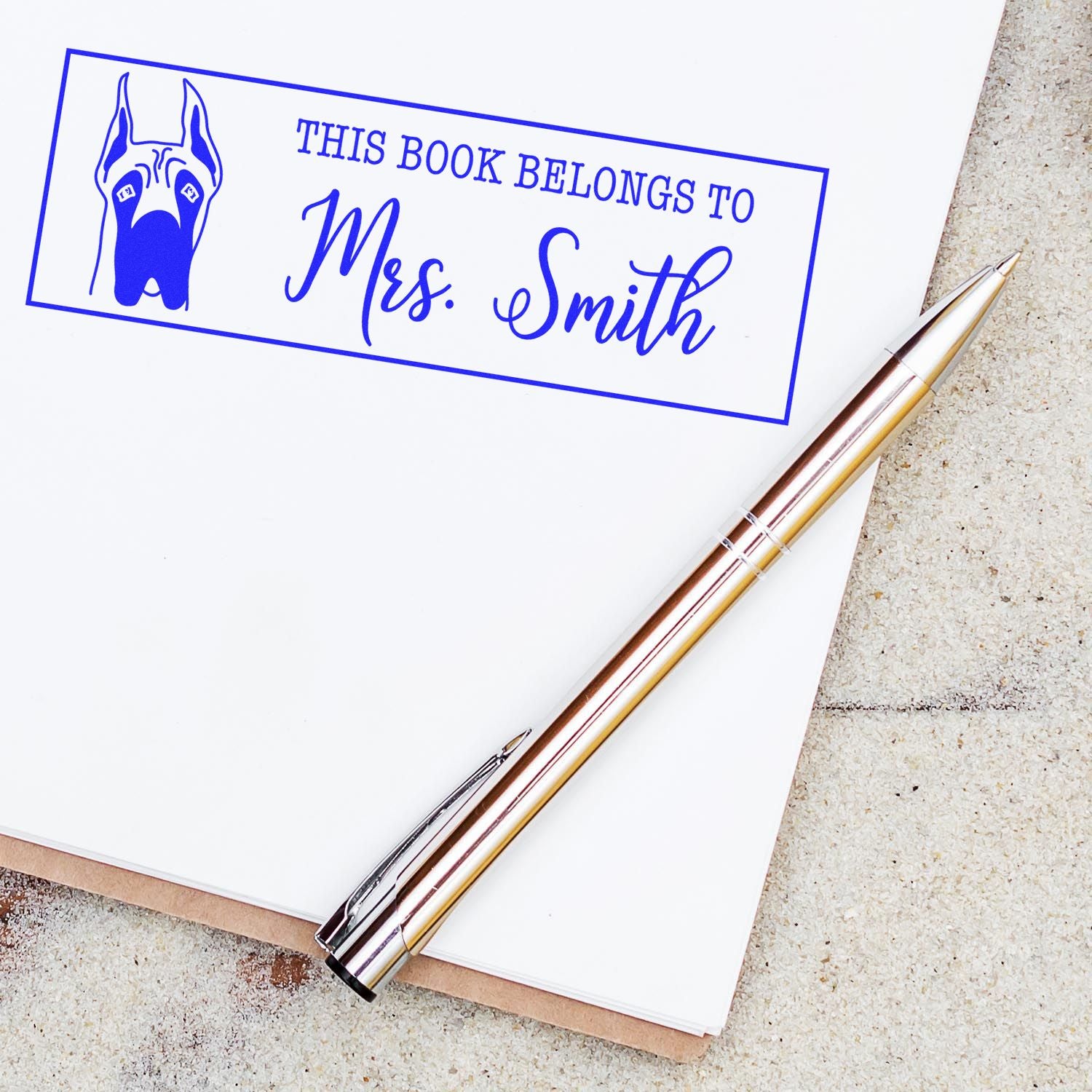 Blue custom book stamp and seal for Mrs. Smith with dog icon, shown on a notebook beside a metallic pen.