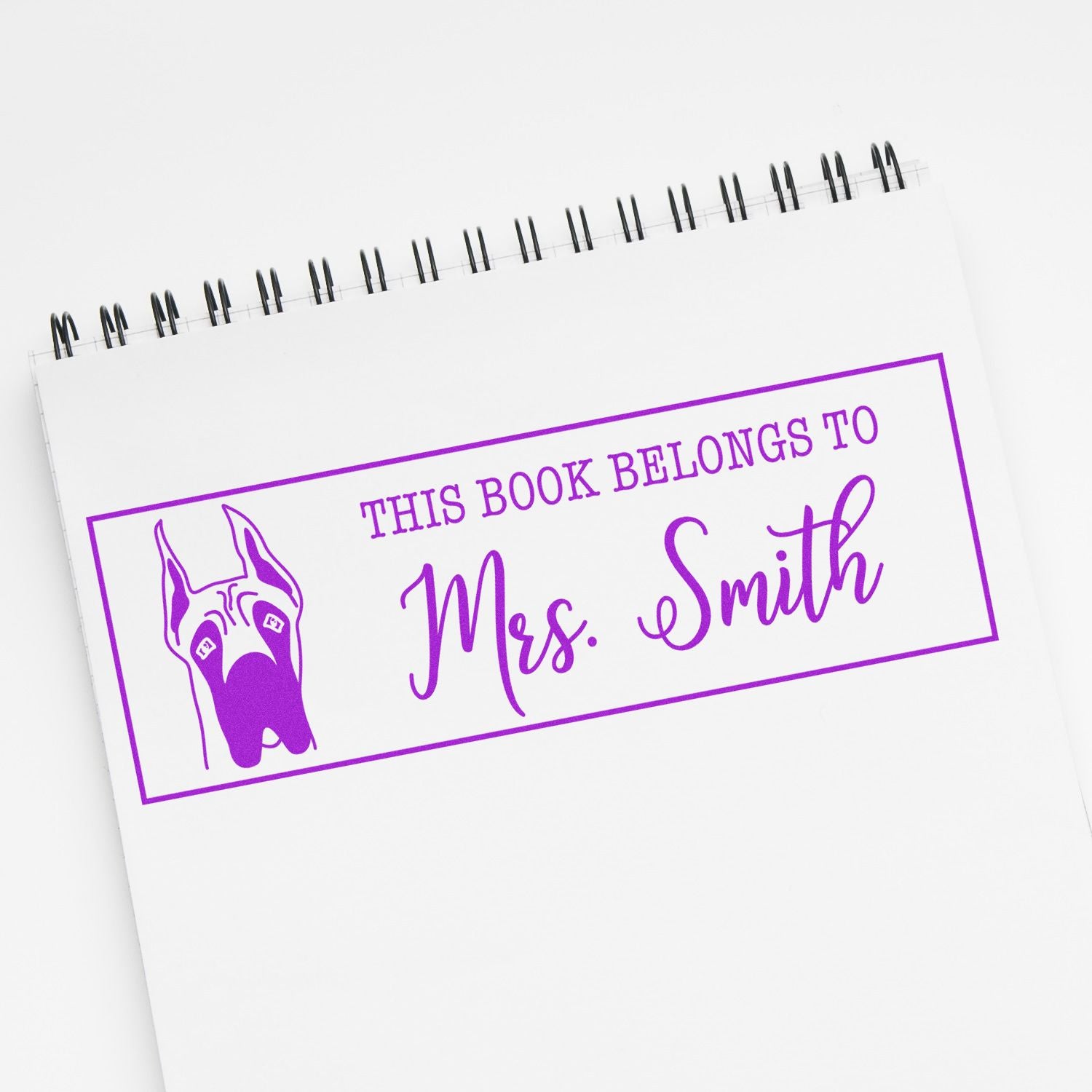Purple custom book stamp with dog seal reading This book belongs to Mrs. Smith on a white spiral notebook.