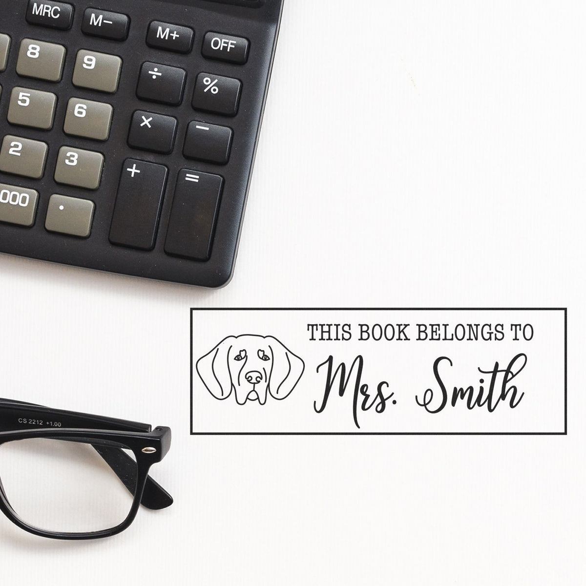 Black book stamp seal with dog outline and This Book Belongs to Mrs. Smith on white, beside a calculator and glasses.