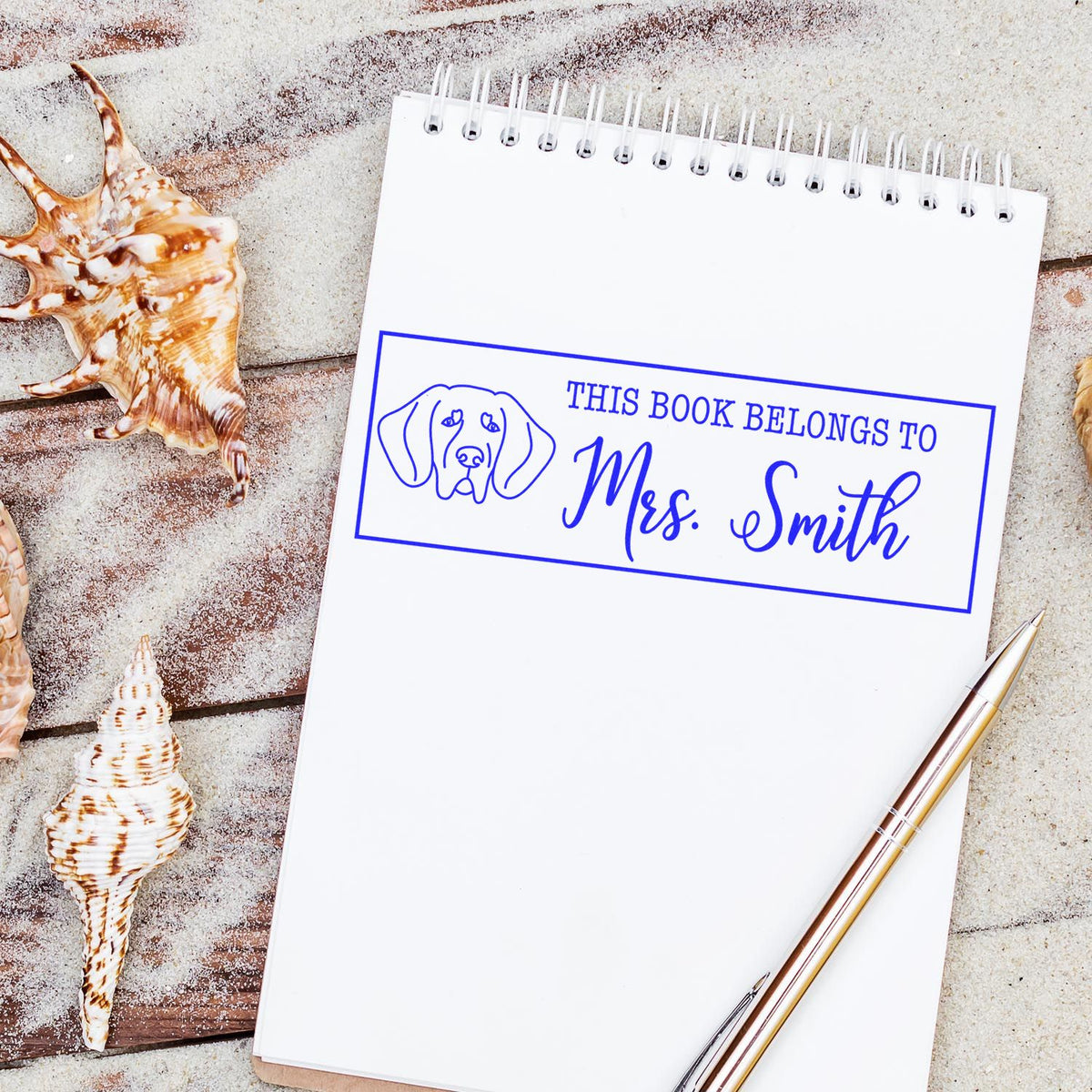 Blue personalized book stamp seal on a white spiral notebook with dog icon, seashells, and a gold pen on a sandy background.