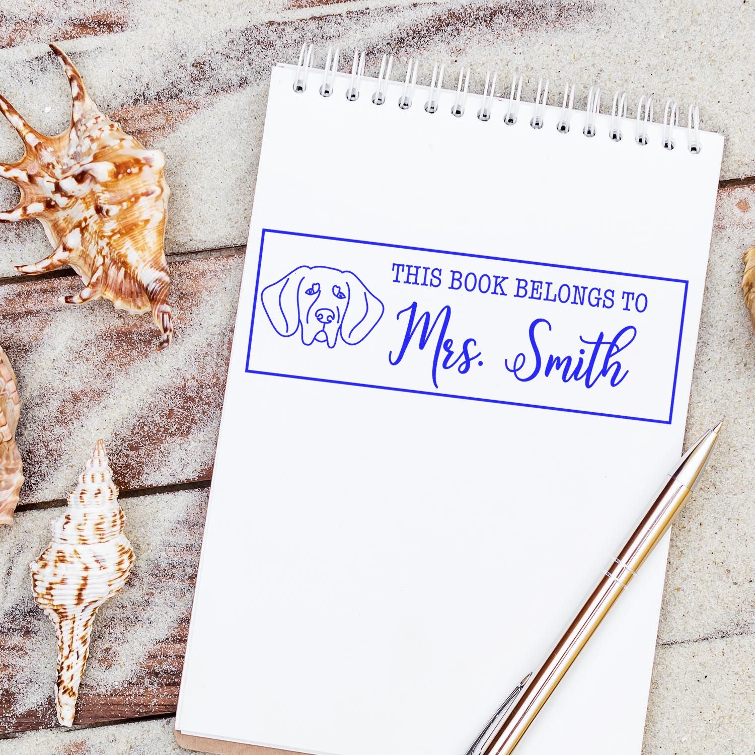 Blue custom book stamp and seal for Mrs. Smith with dog icon on a white notebook, seashells and pen.