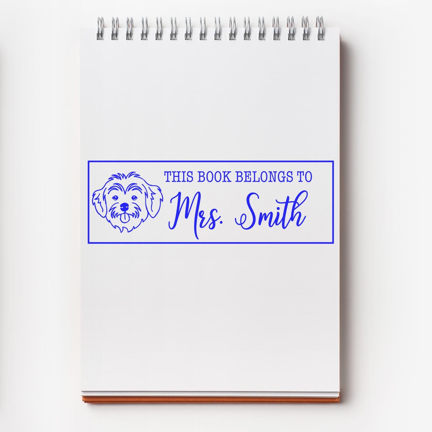 Blue custom book stamp seal with dog illustration and This book belongs to Mrs. Smith on a white spiral notebook.