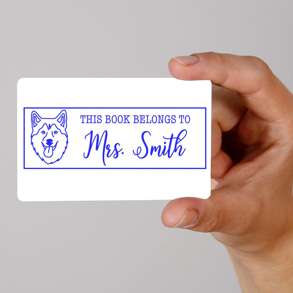 Blue custom book stamp seal with dog icon and This Book Belongs to Mrs. Smith, on a white card, held in hand.