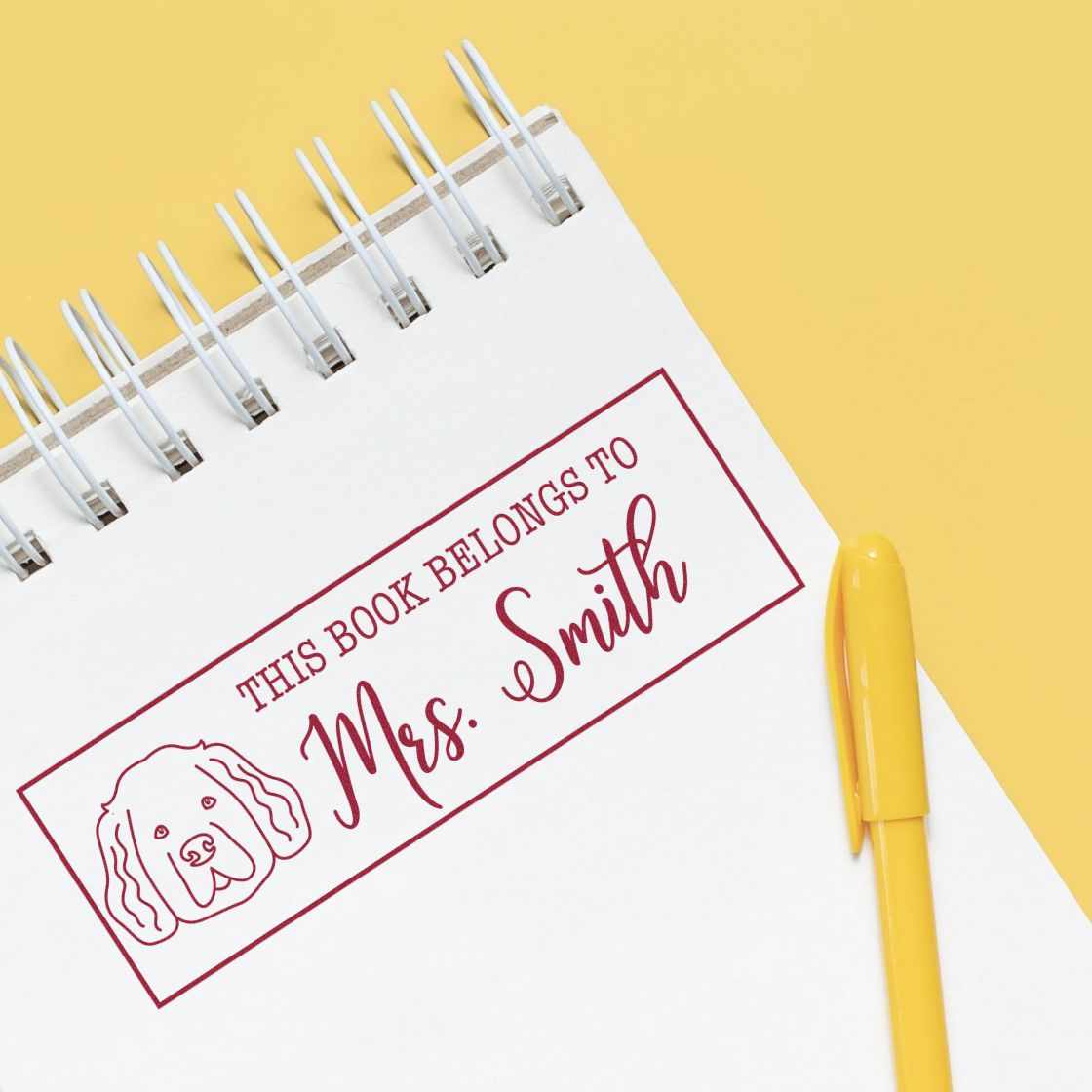 Burgundy personalized book stamp with dog icon and Mrs. Smith text on white spiral notebook; yellow pen on yellow background.