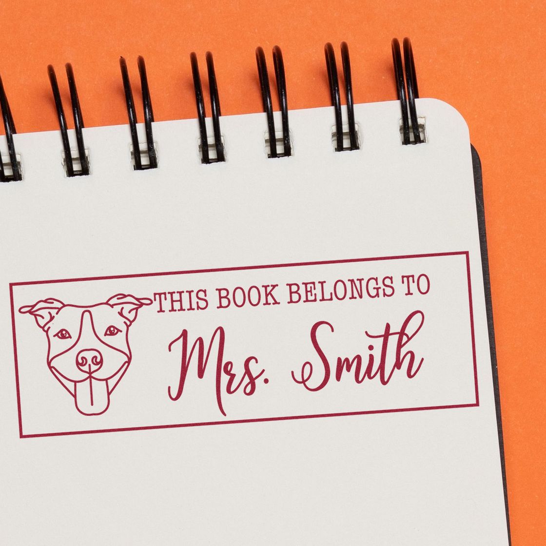 Maroon custom book stamp/seal with dog illustration and This Book Belongs to Mrs. Smith on white spiral notebook over orange background.