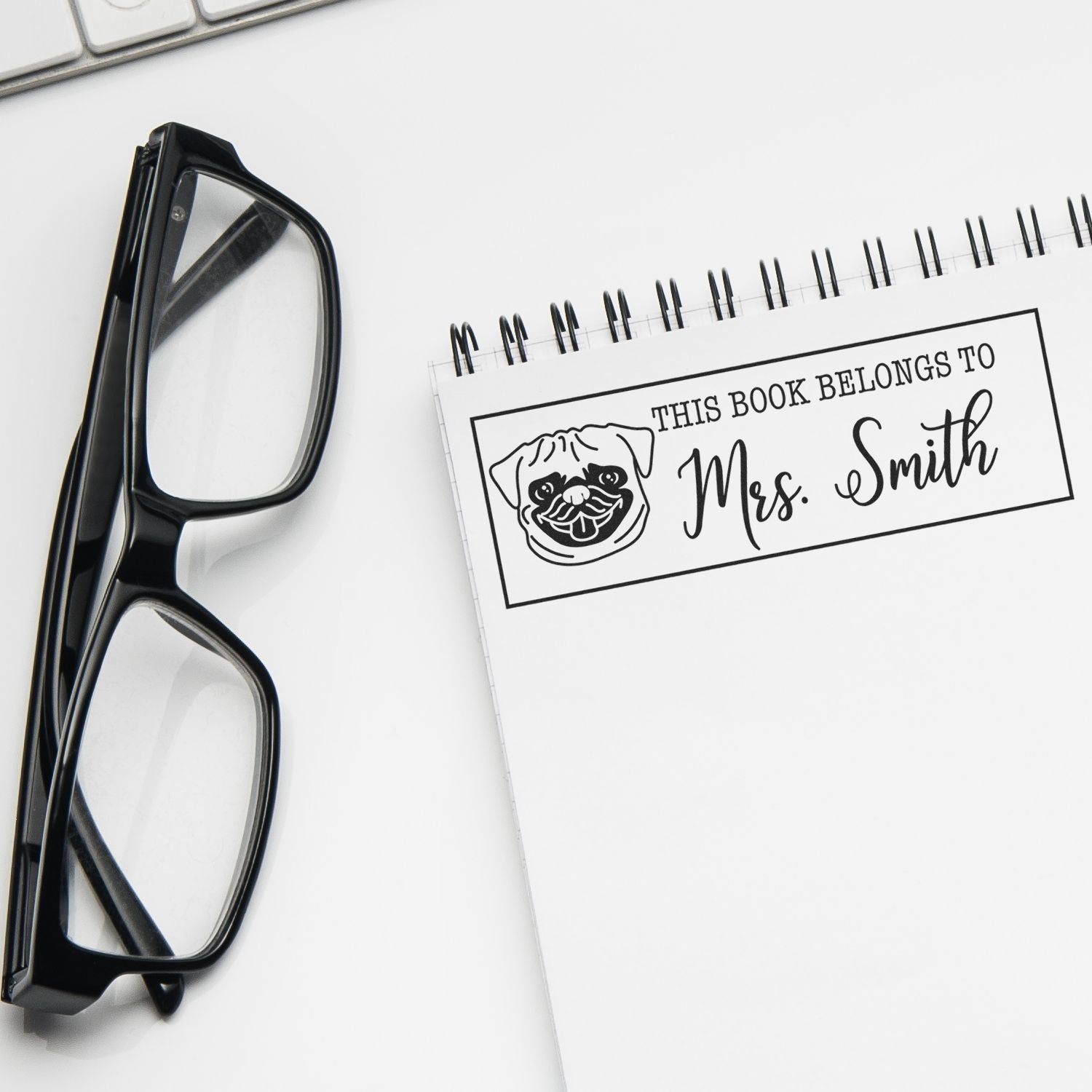 Black eyeglasses beside a white spiral notebook with a custom dog bookplate stamp and seal reading This Book Belongs to Mrs. Smith.