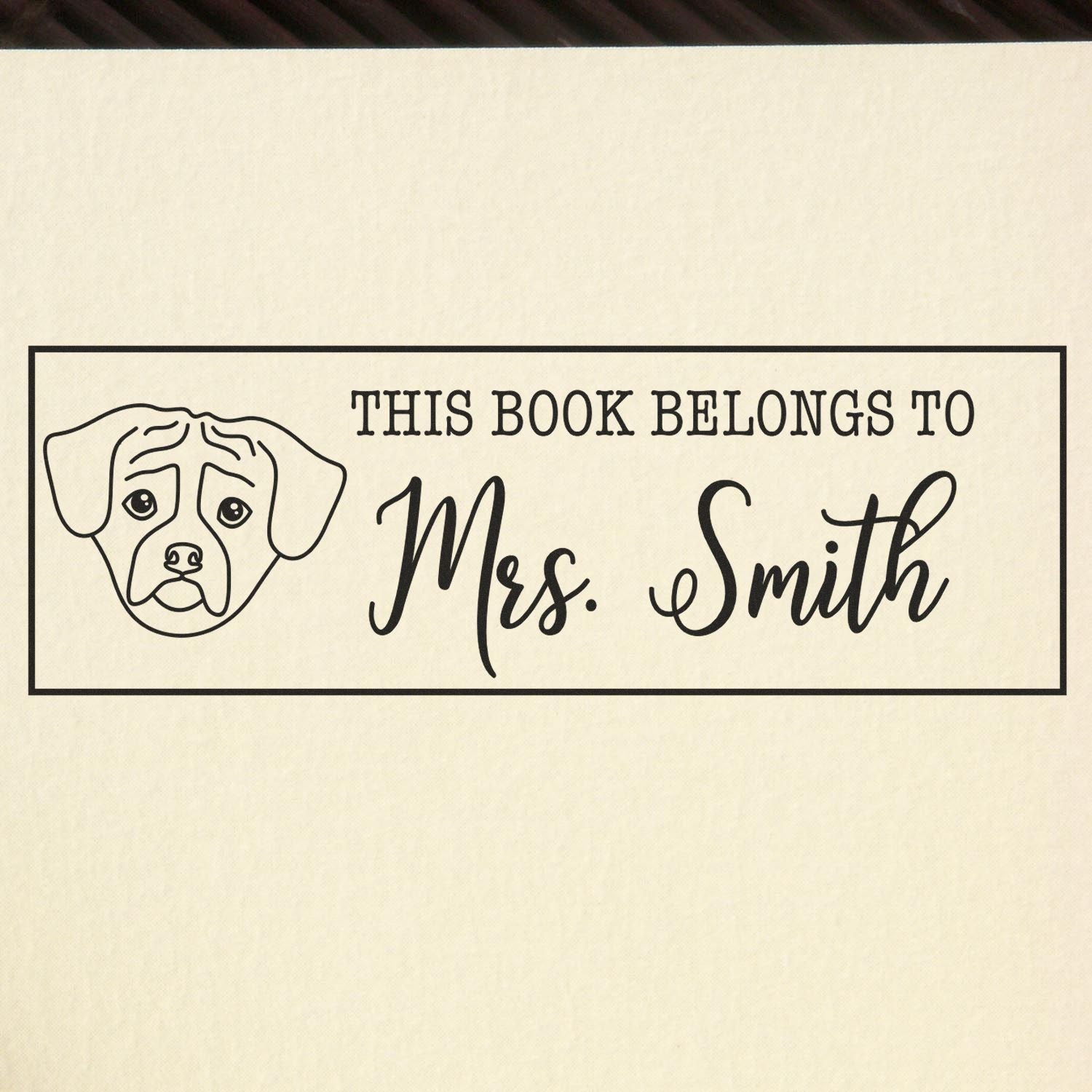 Black custom book stamp with dog face and This Book Belongs to Mrs. Smith on cream paper background.