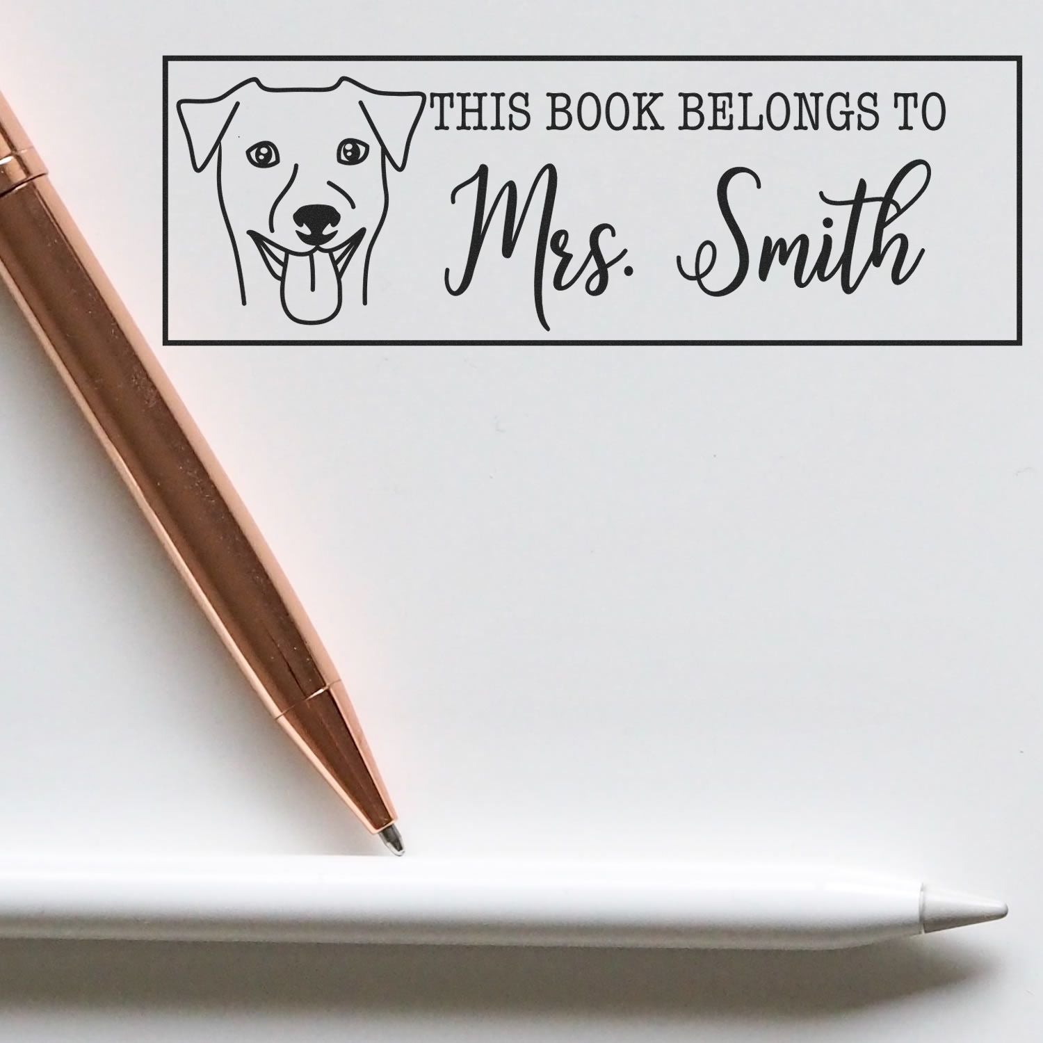 Black custom book stamp with dog illustration and This Book Belongs to Mrs. Smith, shown with rose gold and white pens.