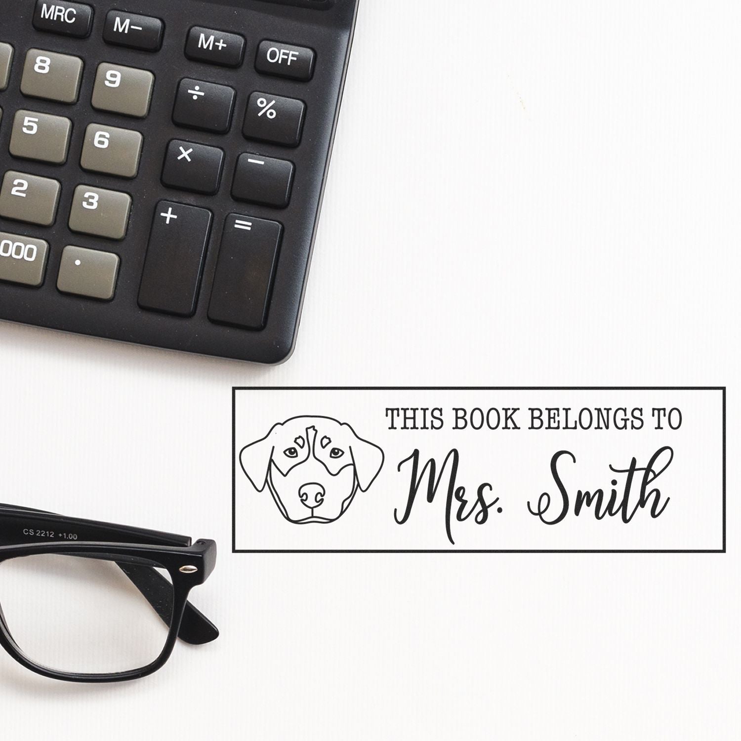 Black personalized book stamp and seal with dog outline and This Book Belongs to Mrs. Smith on white beside calculator and glasses.
