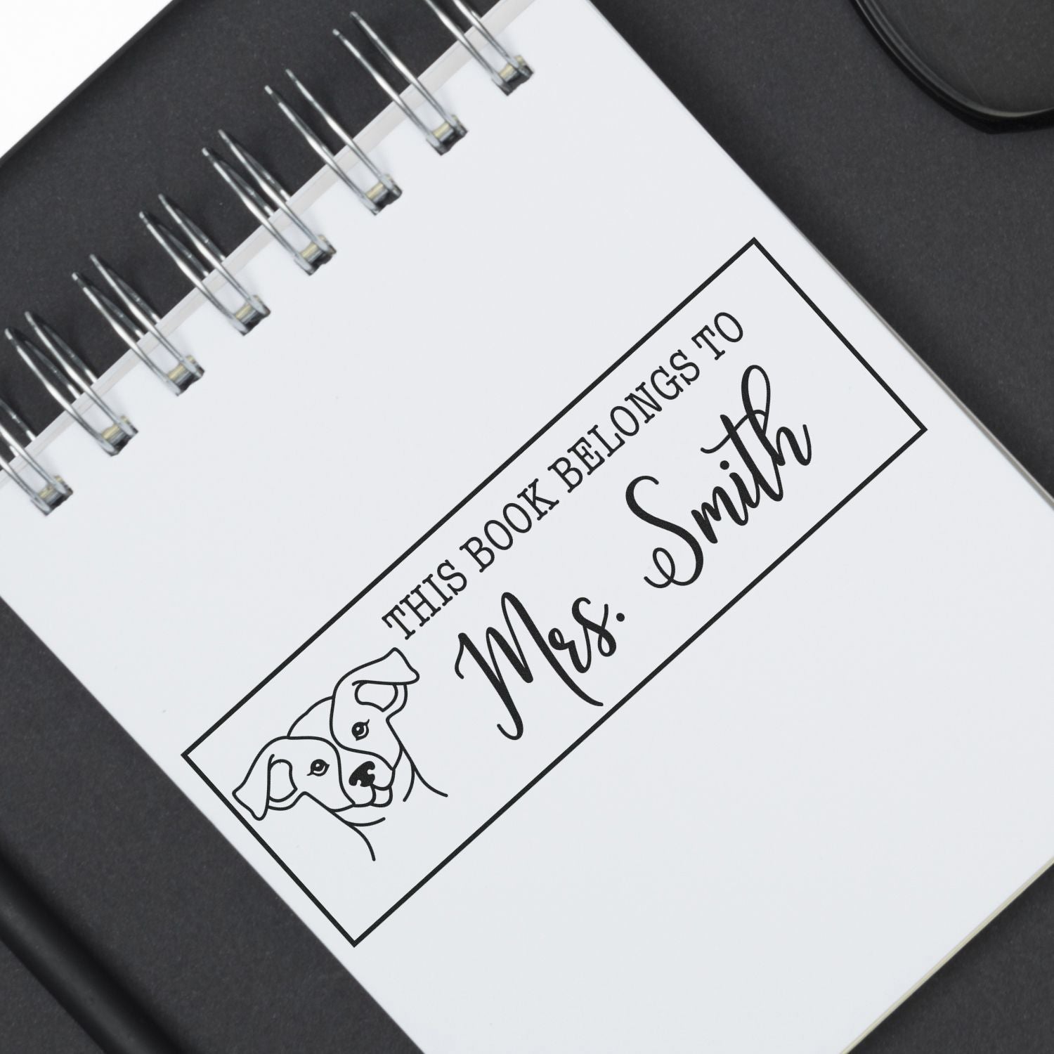 Black custom book stamp seal on a white spiral notebook, reading This Book Belongs to Mrs. Smith with dog line art.