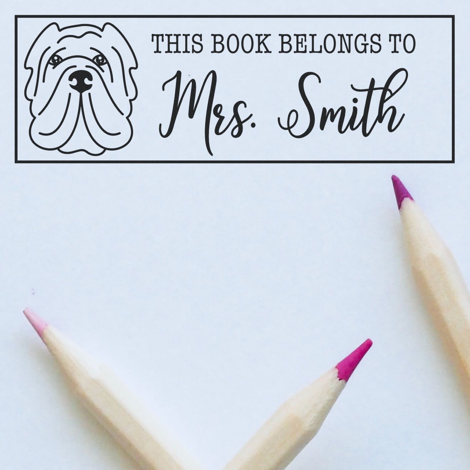 Black custom bookplate stamp seal with bulldog and This Book Belongs to Mrs. Smith on white paper, pink pencils.