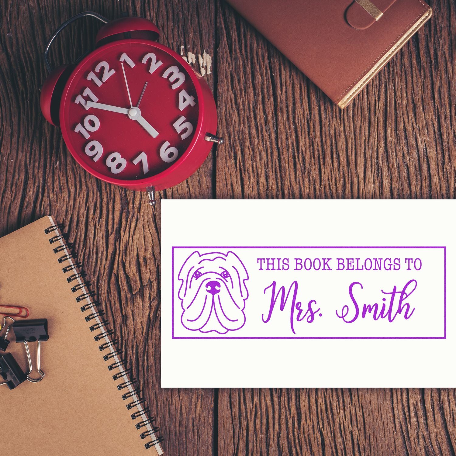 Purple custom book stamp and seal with dog icon on white card, beside a red alarm clock and notebooks on a rustic wood desk.
