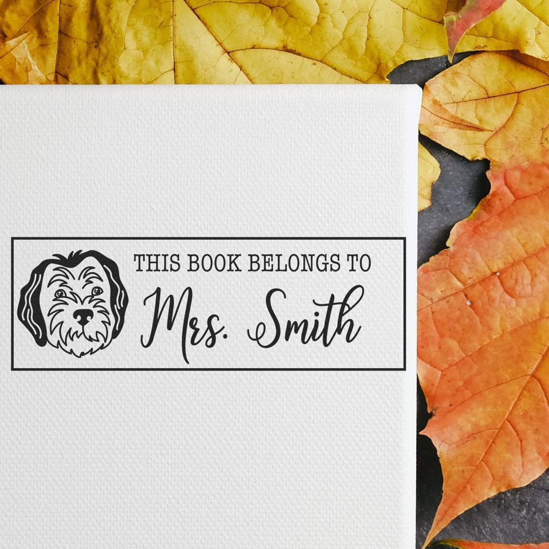 Black custom book stamp reading This Book Belongs to Mrs. Smith with dog icon on white paper and autumn leaves.