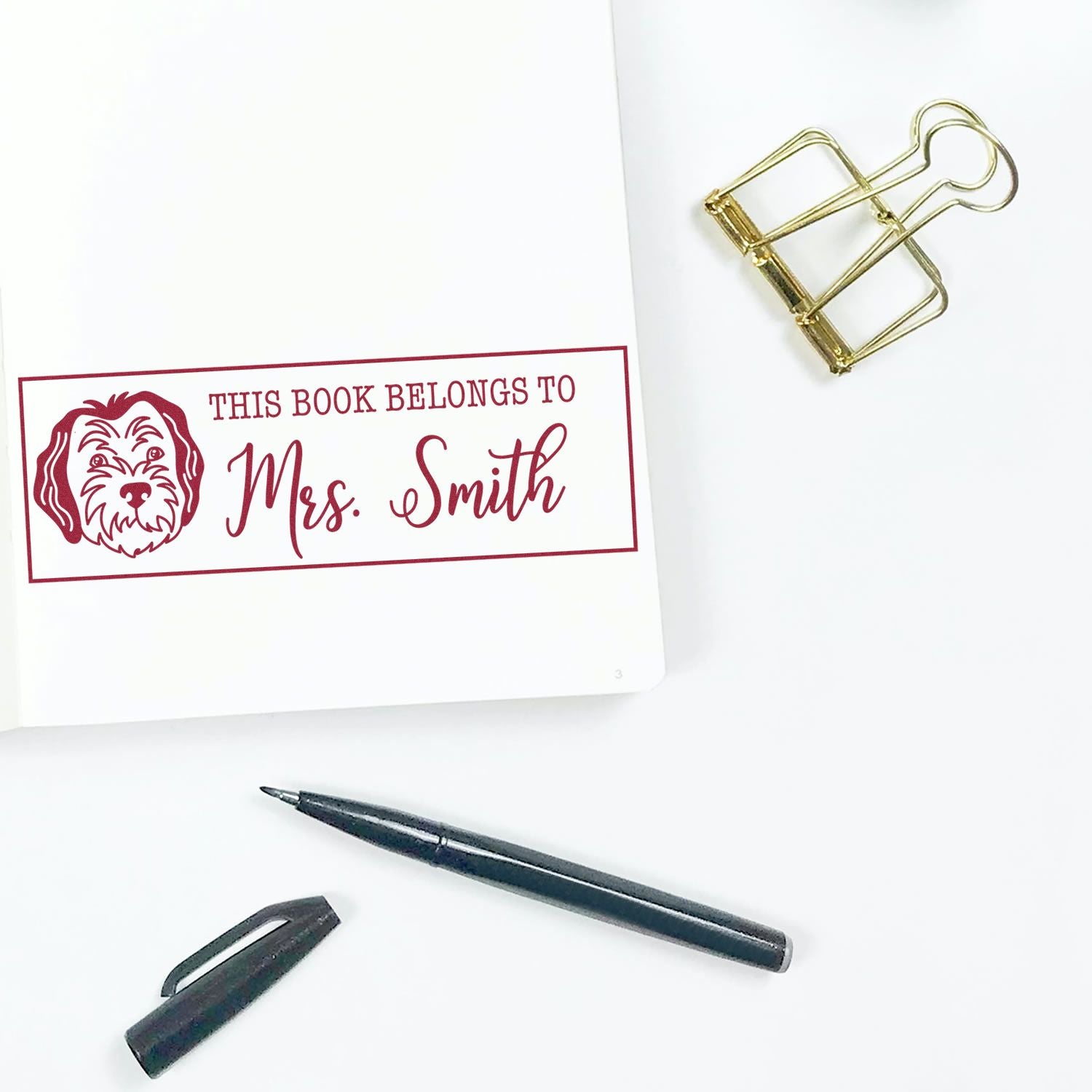 Red personalized book stamp reading This Book Belongs to Mrs. Smith with dog icon, gold clip, and black pen on white.