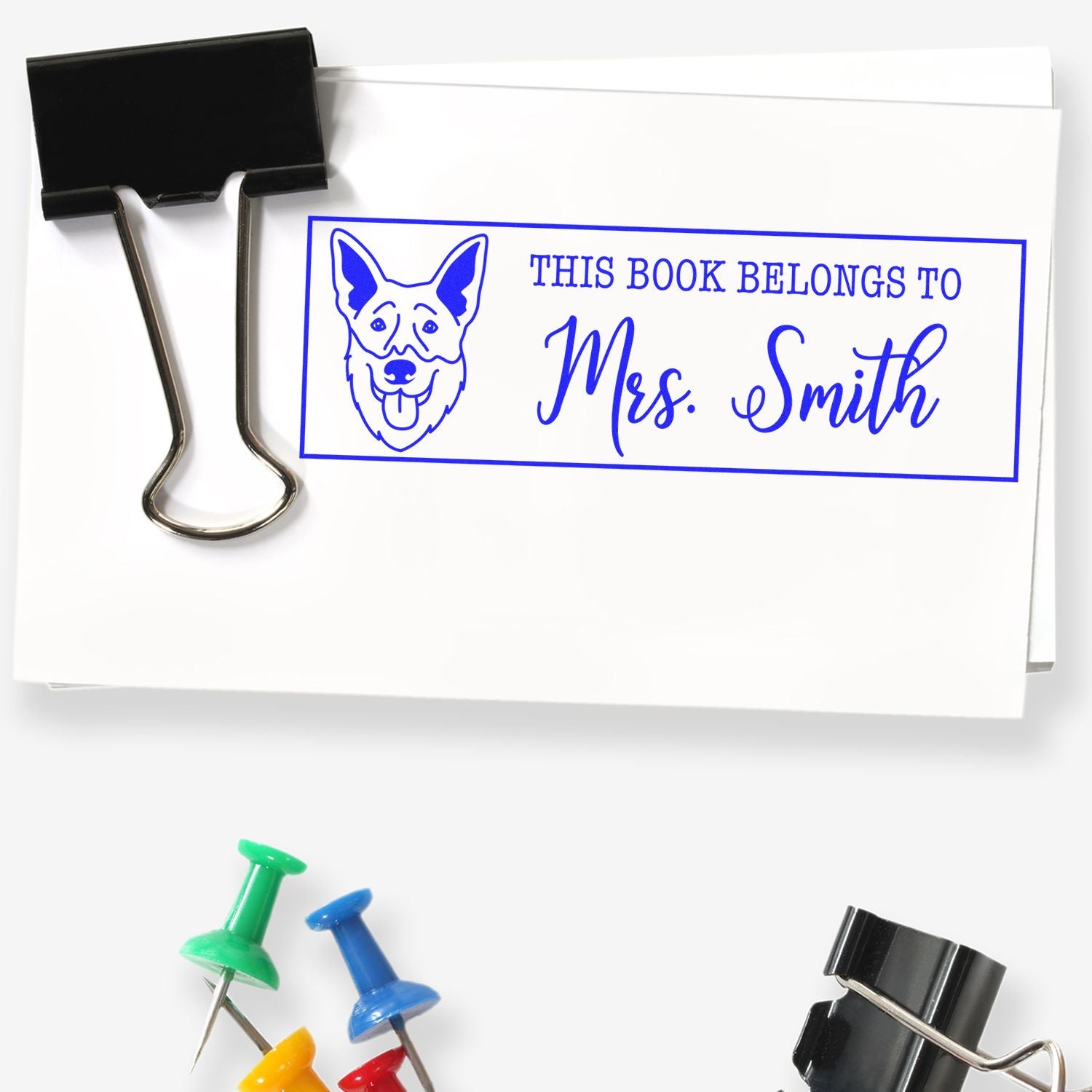 Black binder clip holds white paper with a blue personalized book stamp and dog seal reading This Book Belongs to Mrs. Smith.
