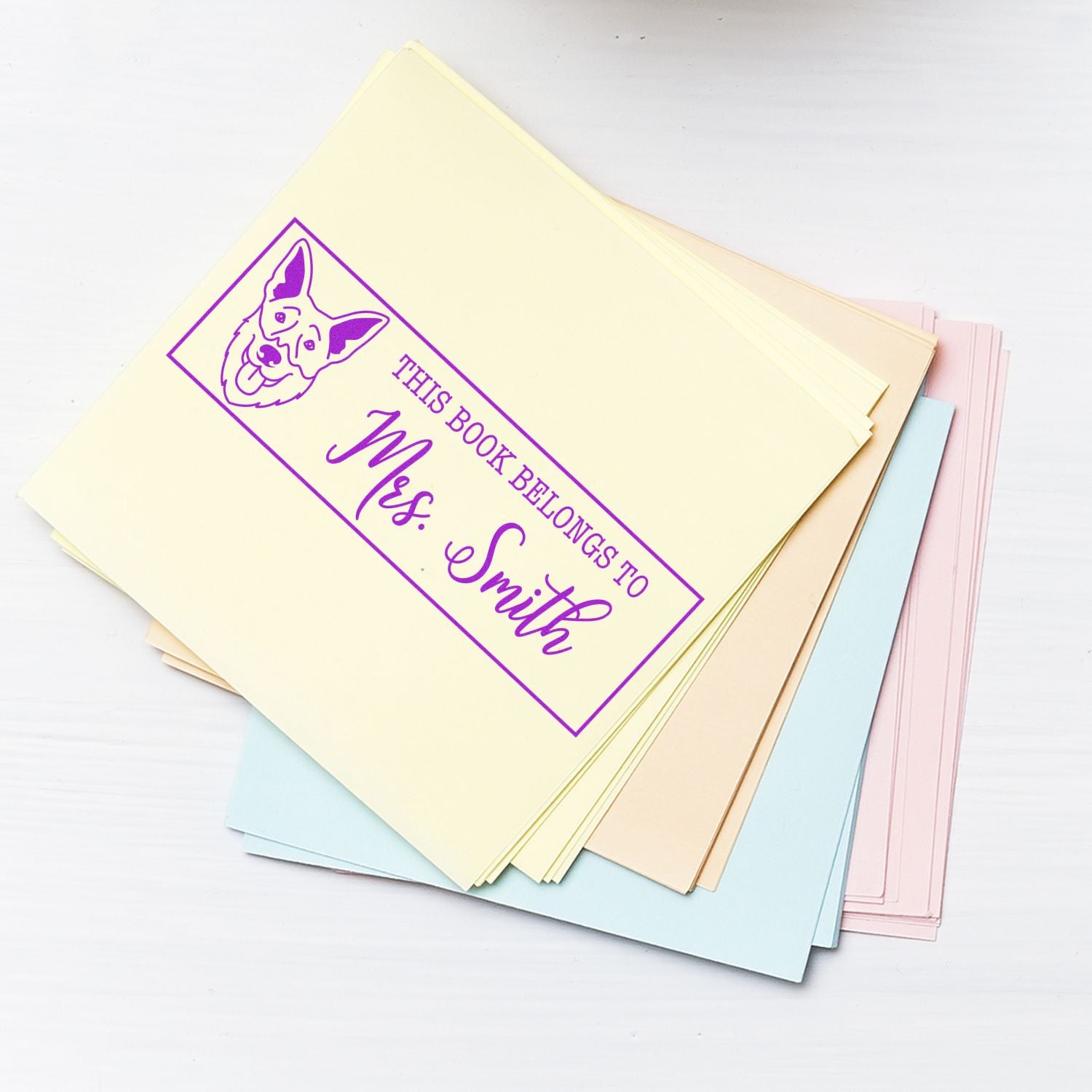 Purple custom book stamp seal with dog illustration and "This book belongs to Mrs. Smith" on pastel paper stack.