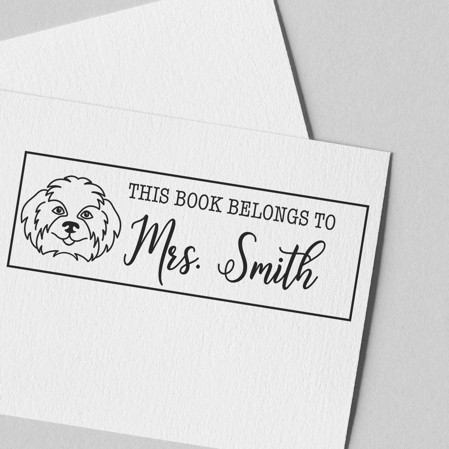 Black personalized book stamp seal with dog illustration and This book belongs to Mrs. Smith on white textured paper.