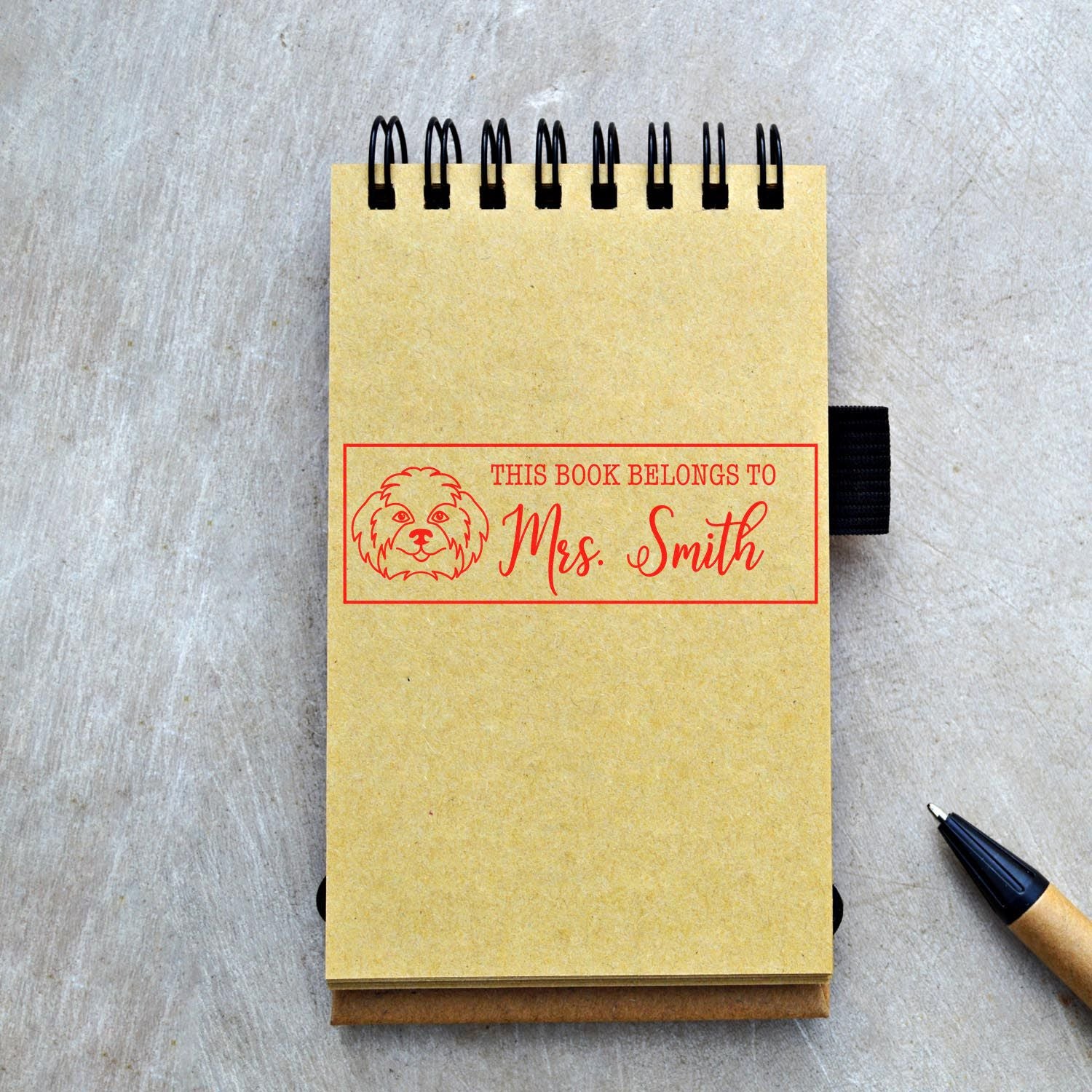 Yellow spiral notebook with red custom book stamp and seal reading This Book Belongs to Mrs. Smith, plus dog icon, on gray table.
