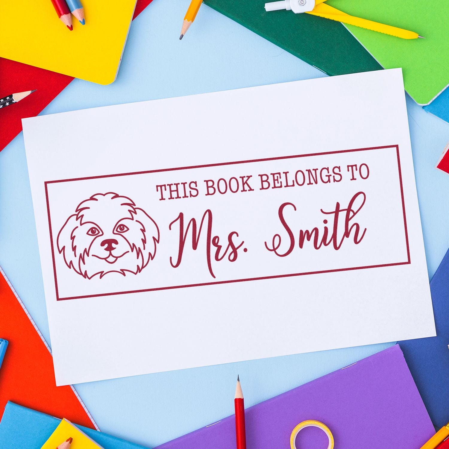 Red custom book stamp/seal reading This Book Belongs to Mrs. Smith with dog icon on white card, surrounded by colorful pencils.