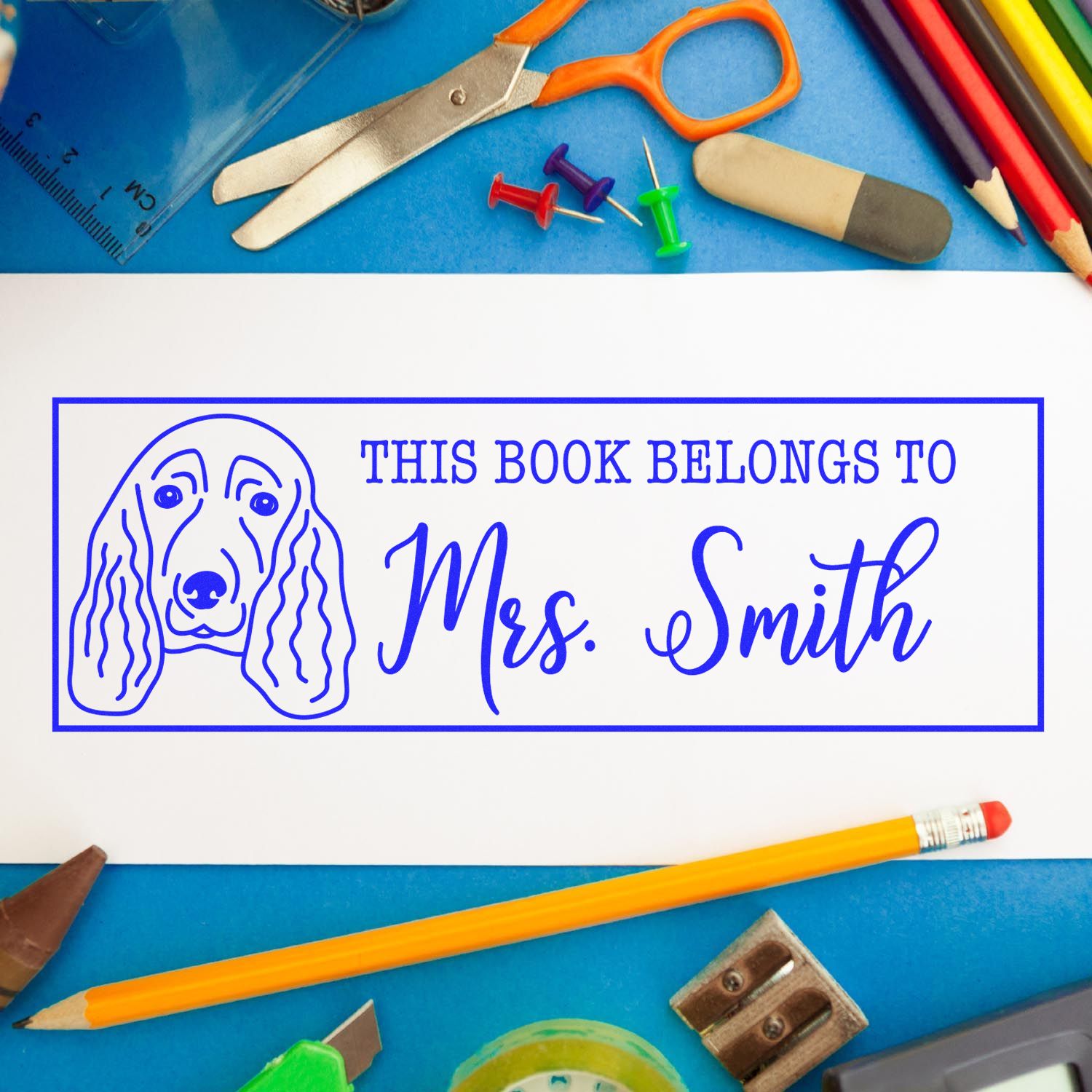 Blue custom book stamp and seal with dog outline reading This Book Belongs to Mrs. Smith, surrounded by school supplies.