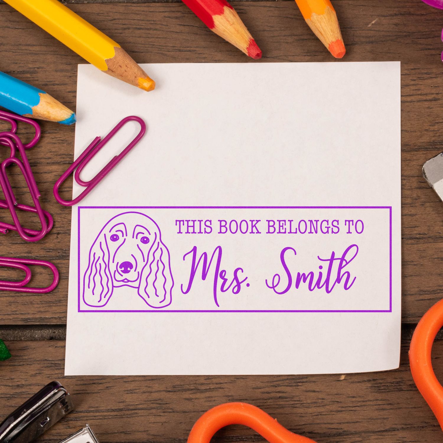 Purple personalized book stamp and seal with dog outline and Mrs. Smith on white paper, surrounded by colorful pencils and paper clips.