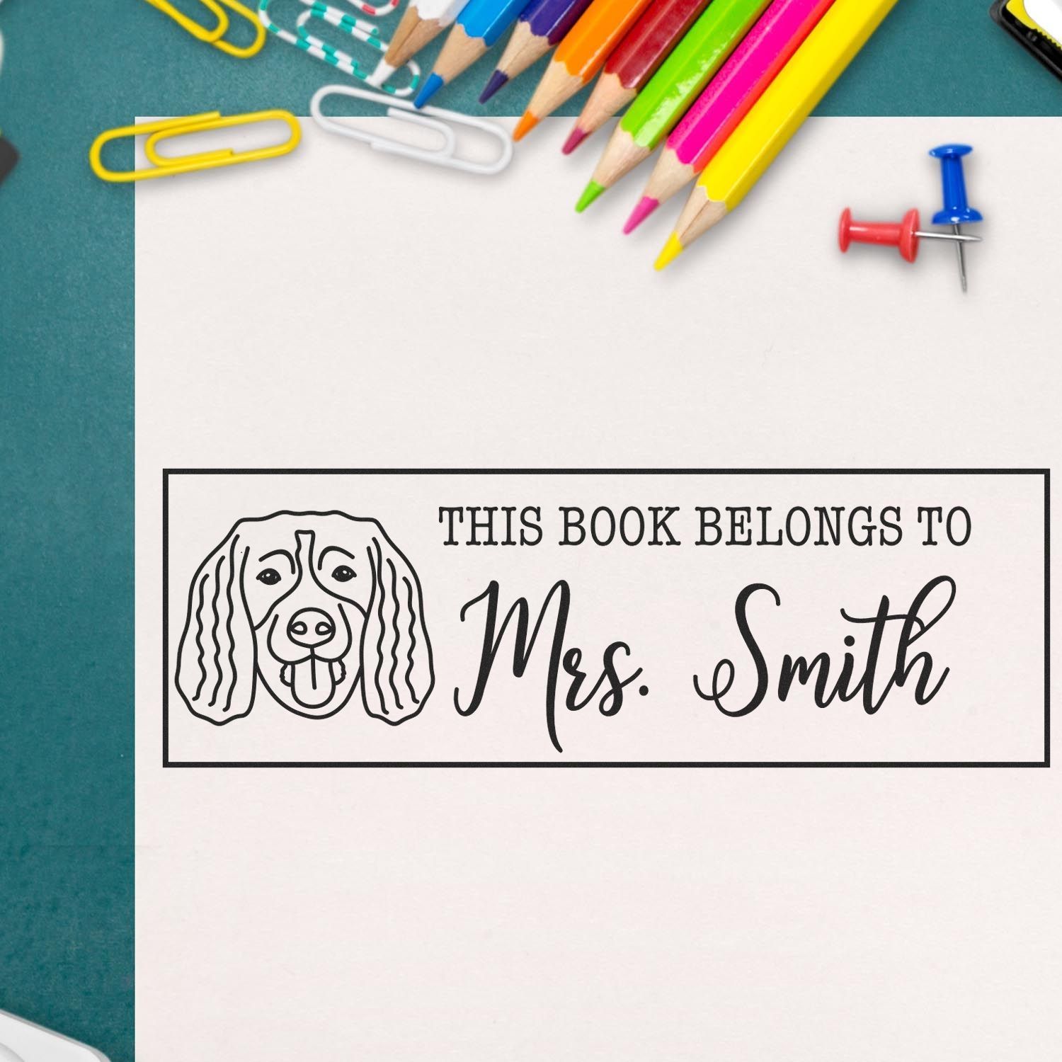 Black personalized bookplate stamp/seal with dog illustration and This Book Belongs to Mrs. Smith on white paper, colorful pencils.