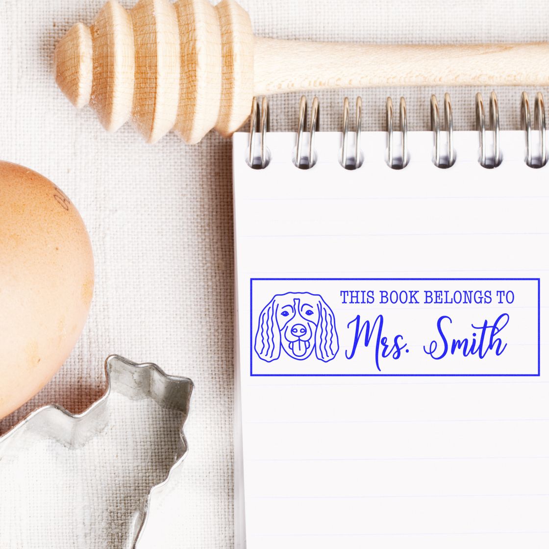 Blue book ownership stamp reading This book belongs to Mrs. Smith on a white notebook, with a wooden seal and egg nearby.
