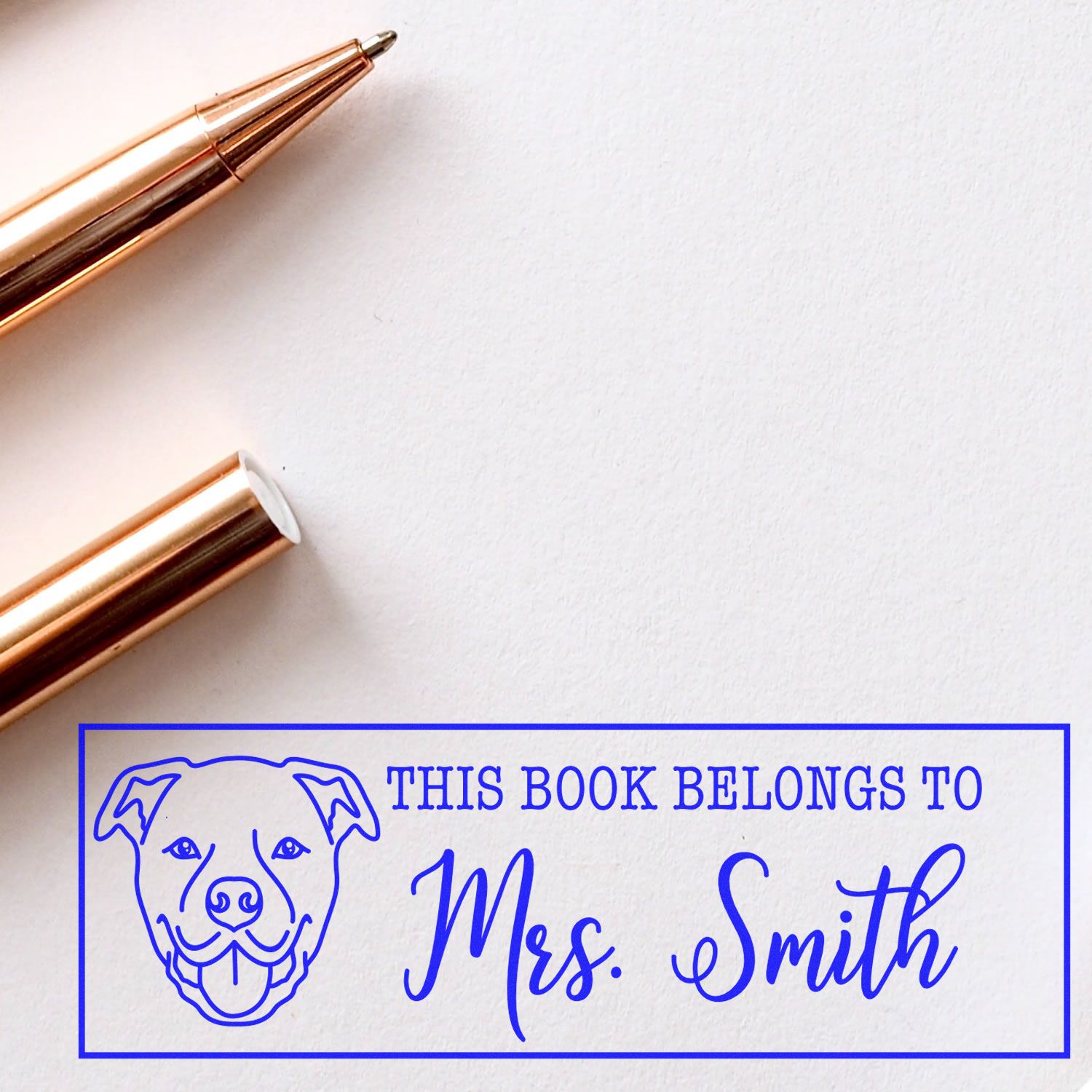 Rose gold wax seal stamper and pen on white paper beside a blue custom book stamp reading This Book Belongs to Mrs. Smith with dog outline.