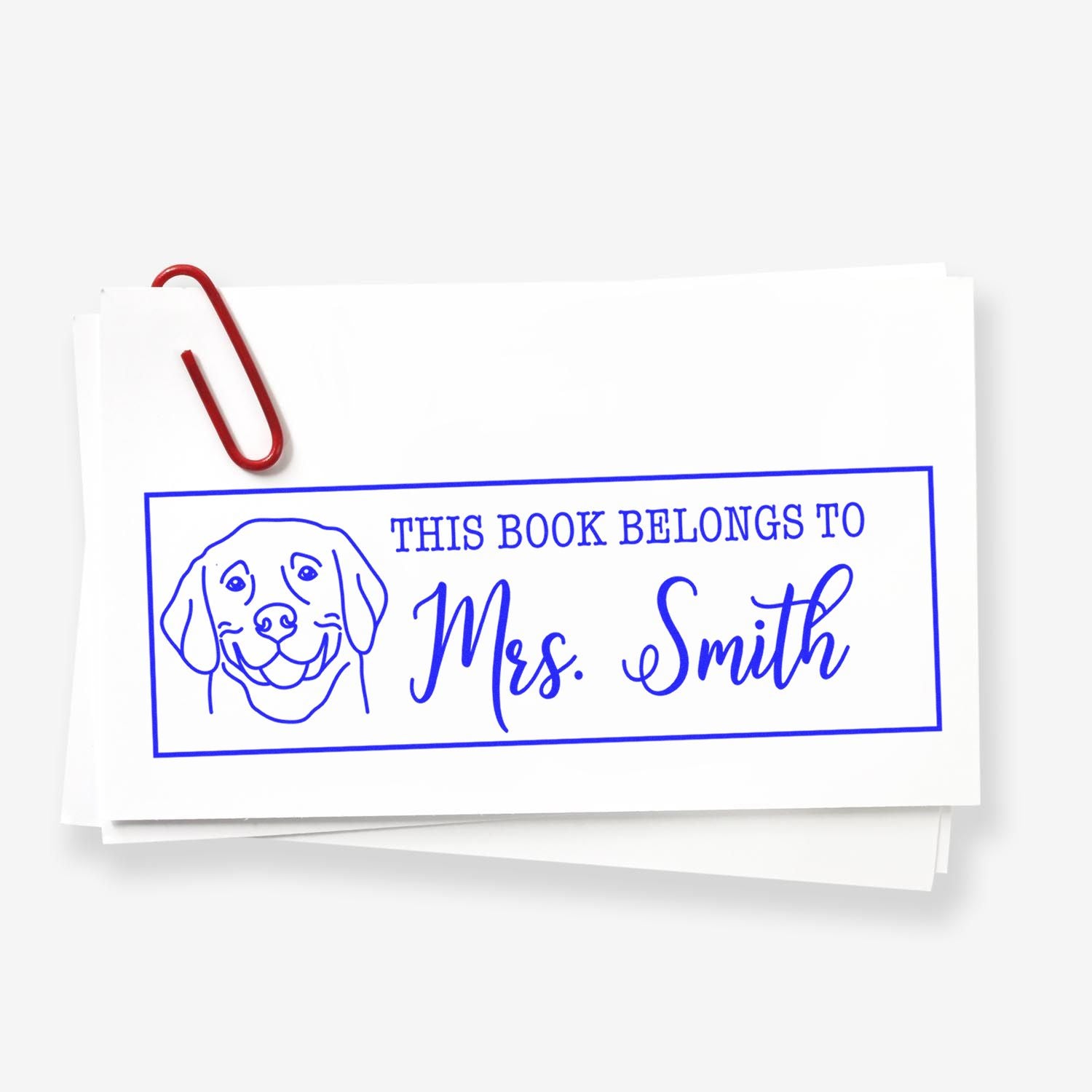 Blue custom book stamp with dog line art and This Book Belongs to Mrs. Smith, clipped with a red paperclip on white paper.