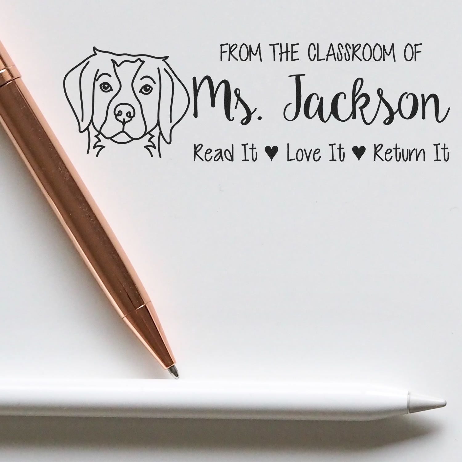 Custom classroom stamp/seal with black dog illustration and Ms. Jackson text, shown with a copper rose-gold pen on white.