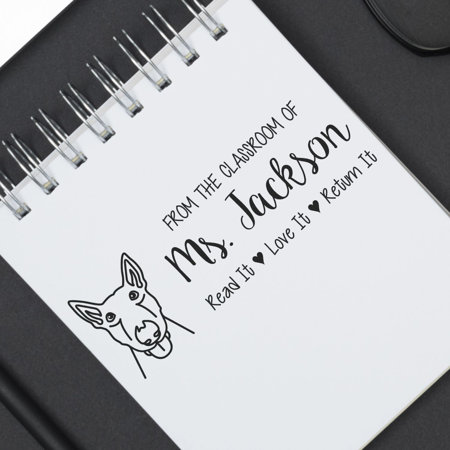 Black custom stamp and seal design on white spiral notebook with dog outline and return-it classroom message.