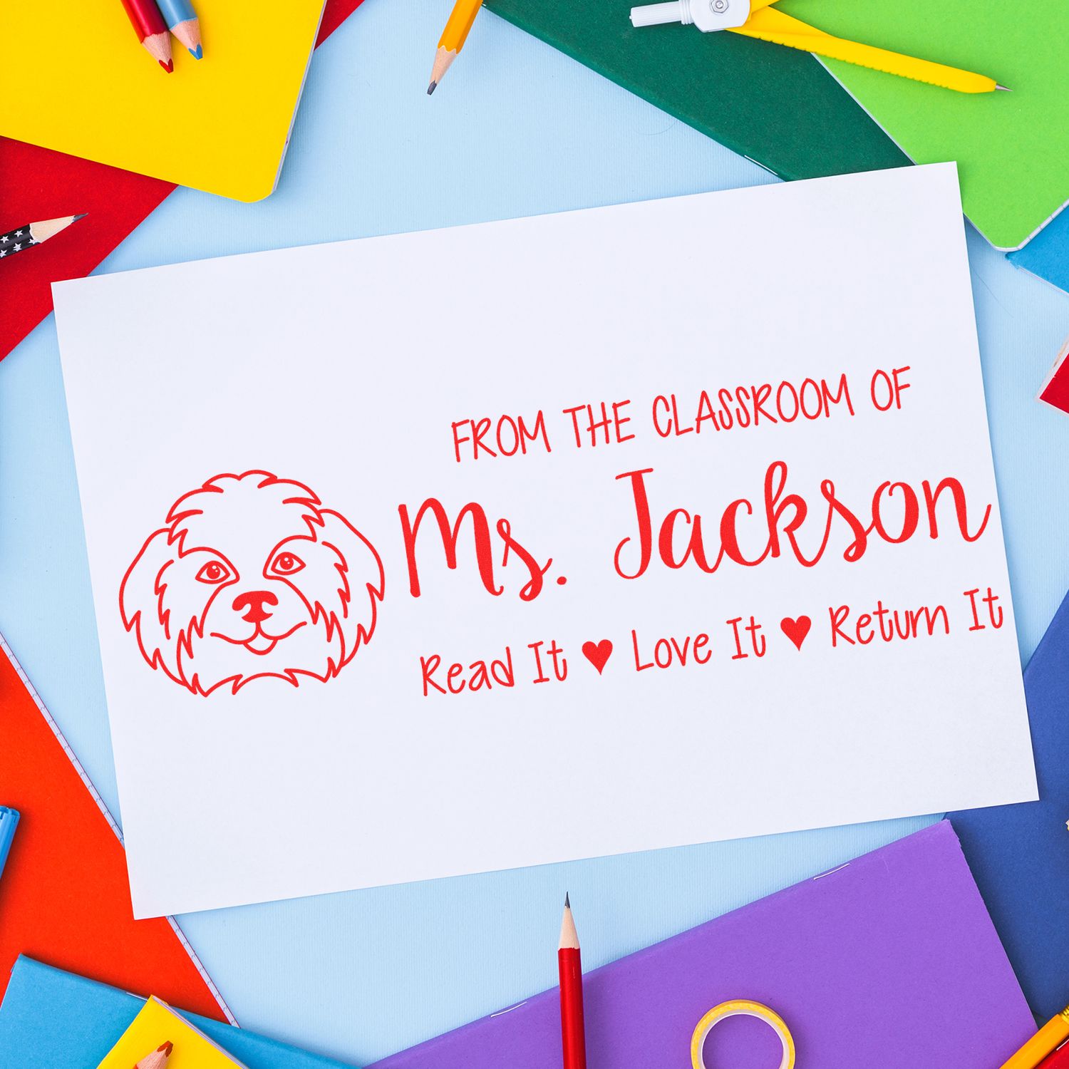 Red custom classroom book stamp with dog logo and Ms. Jackson Read It Love It Return It on white paper, with colorful pencils.