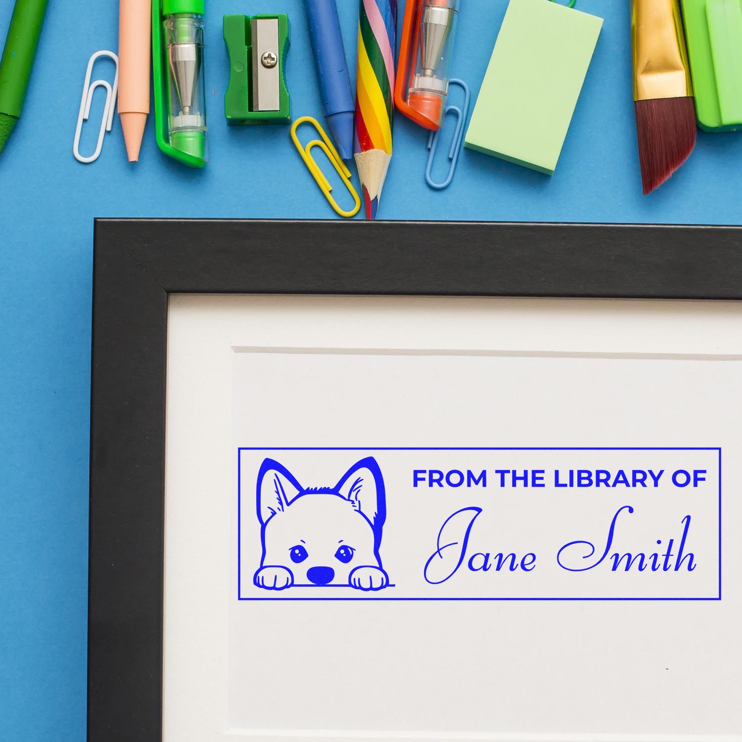 Custom blue library stamp seal with dog icon and Jane Smith on framed print, surrounded by colorful stationery on blue background