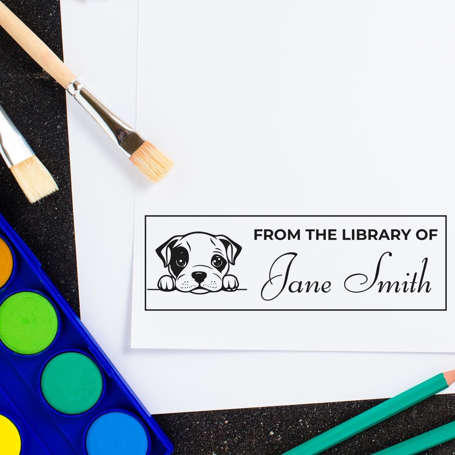 Personalized black library stamp and seal with puppy logo and Jane Smith text on white paper, beside bright watercolor paints.