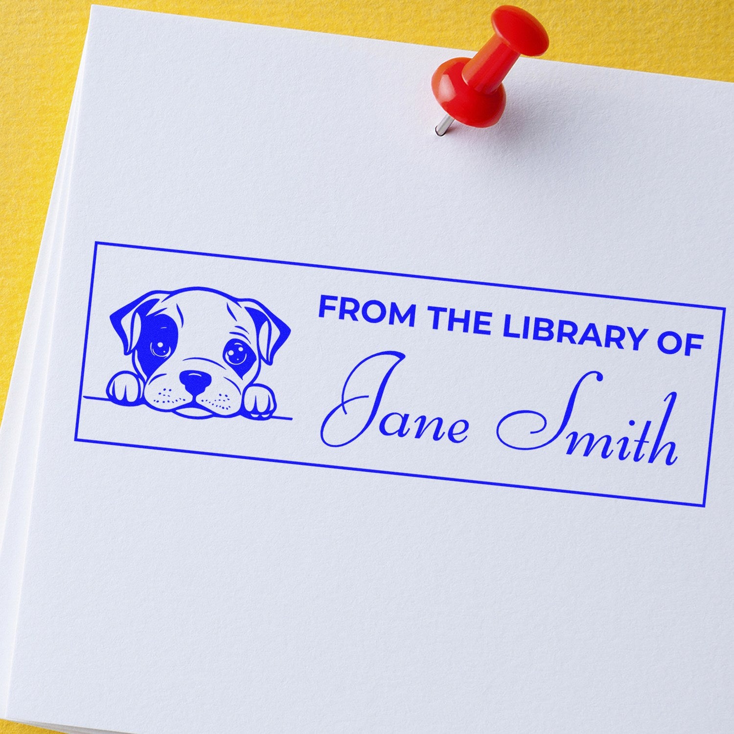 Blue custom library book stamp with puppy seal and red pushpin on white paper over a yellow background.