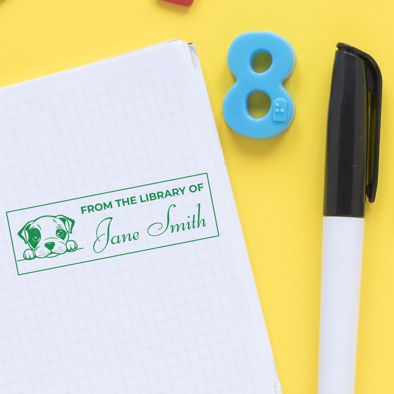 Green custom library stamp with dog and Jane Smith on notebook, beside a blue number 8 seal and black pen on yellow.