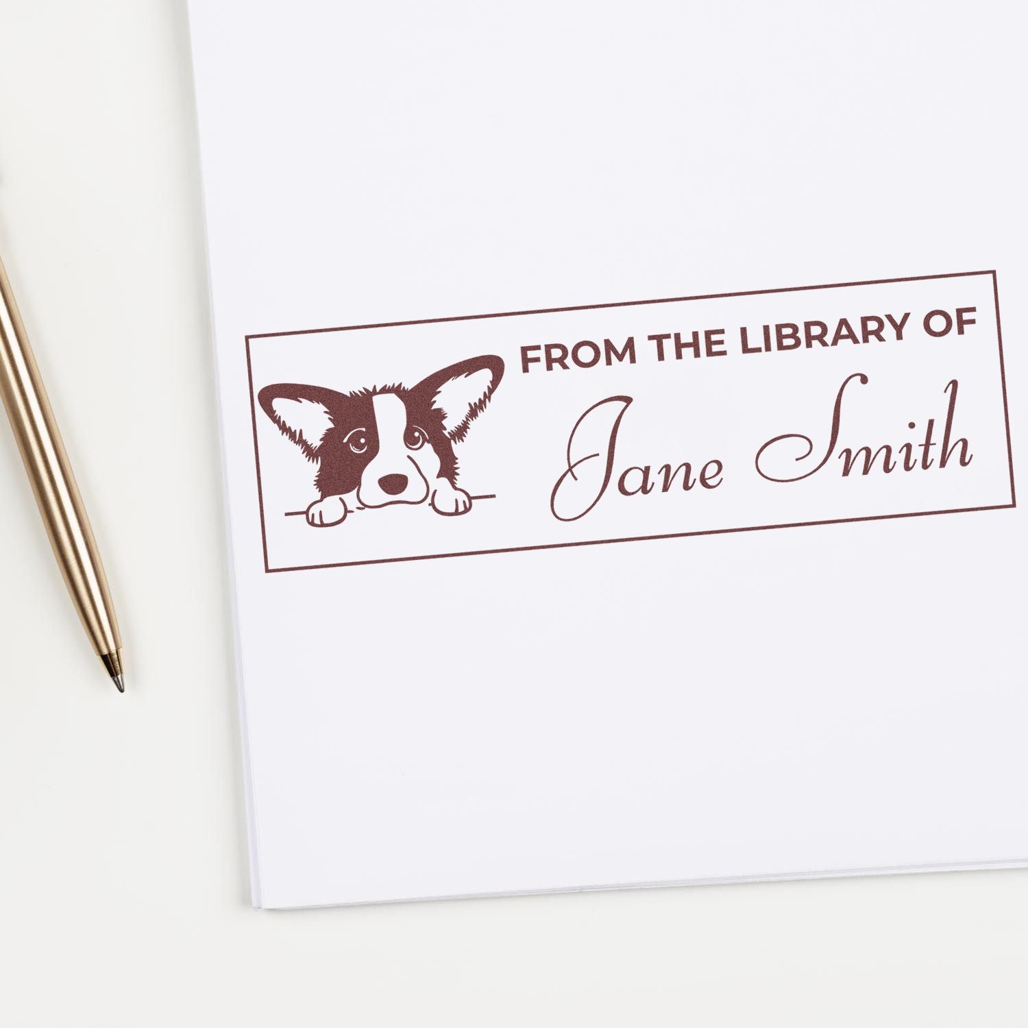 Brown custom library stamp with corgi and From the Library of Jane Smith on white paper beside a gold pen.