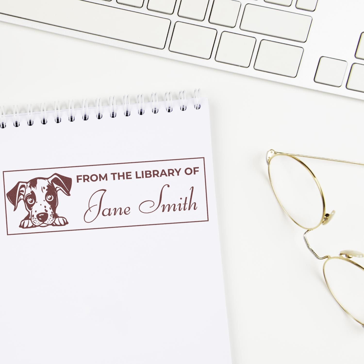 Personalized brown library stamp and seal with dog illustration and Jane Smith on a white notebook, beside gold glasses and keyboard.