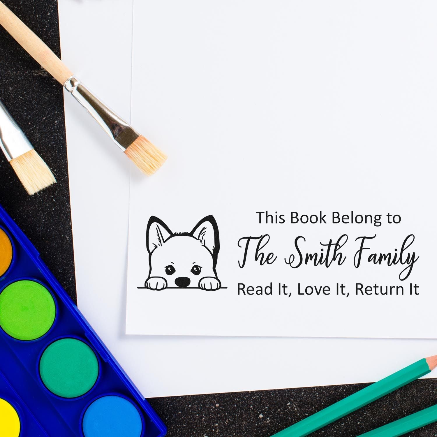 Personalized book stamp and seal with dog graphic on white paper, surrounded by paintbrushes, colored pencils and watercolor paints.
