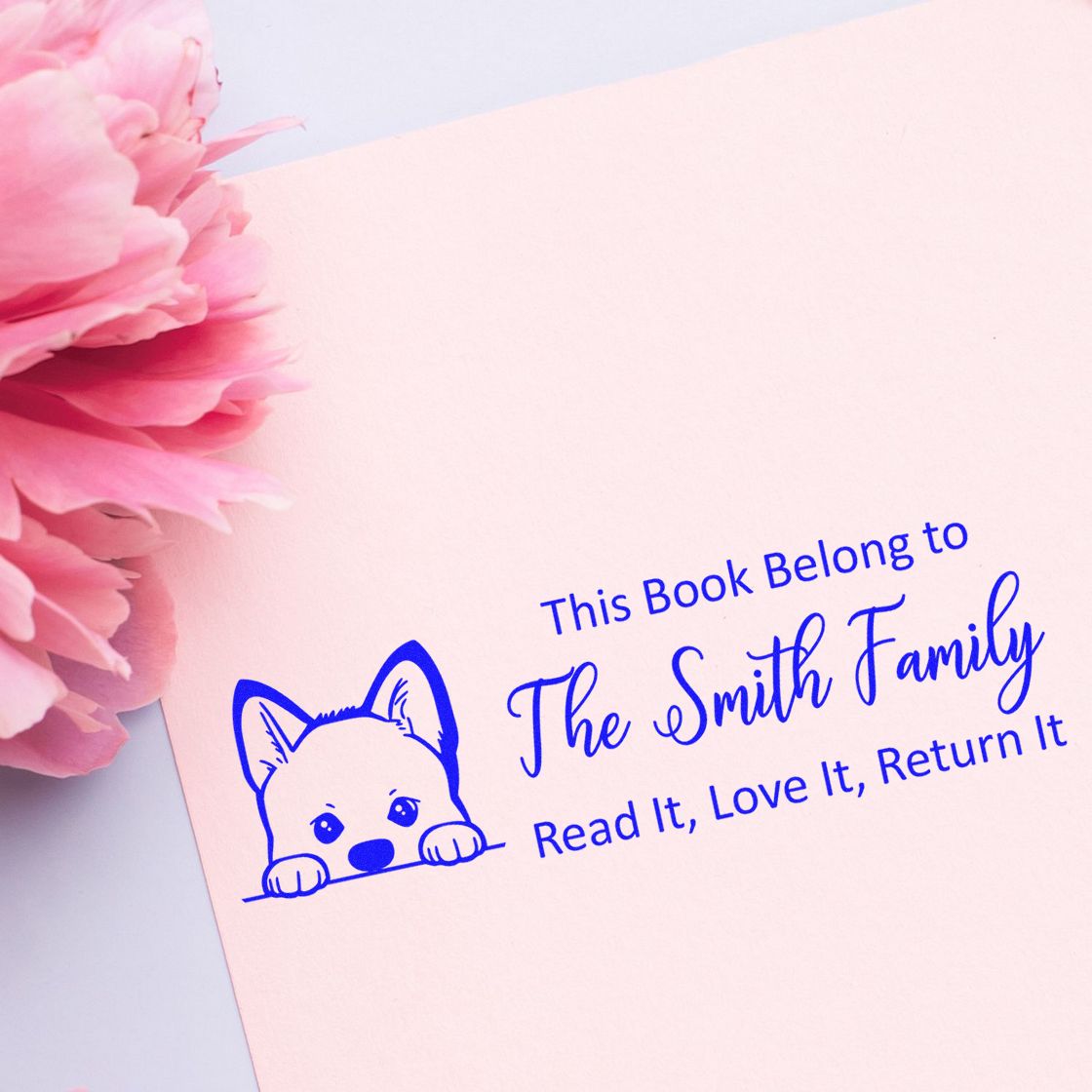 Blue custom book ownership stamp seal with dog icon on pale pink paper, beside pink flowers.