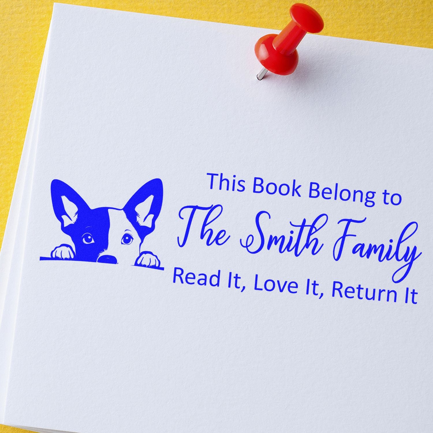 Blue custom book stamp seal with peeking dog logo and family name on white paper, held by red push pin